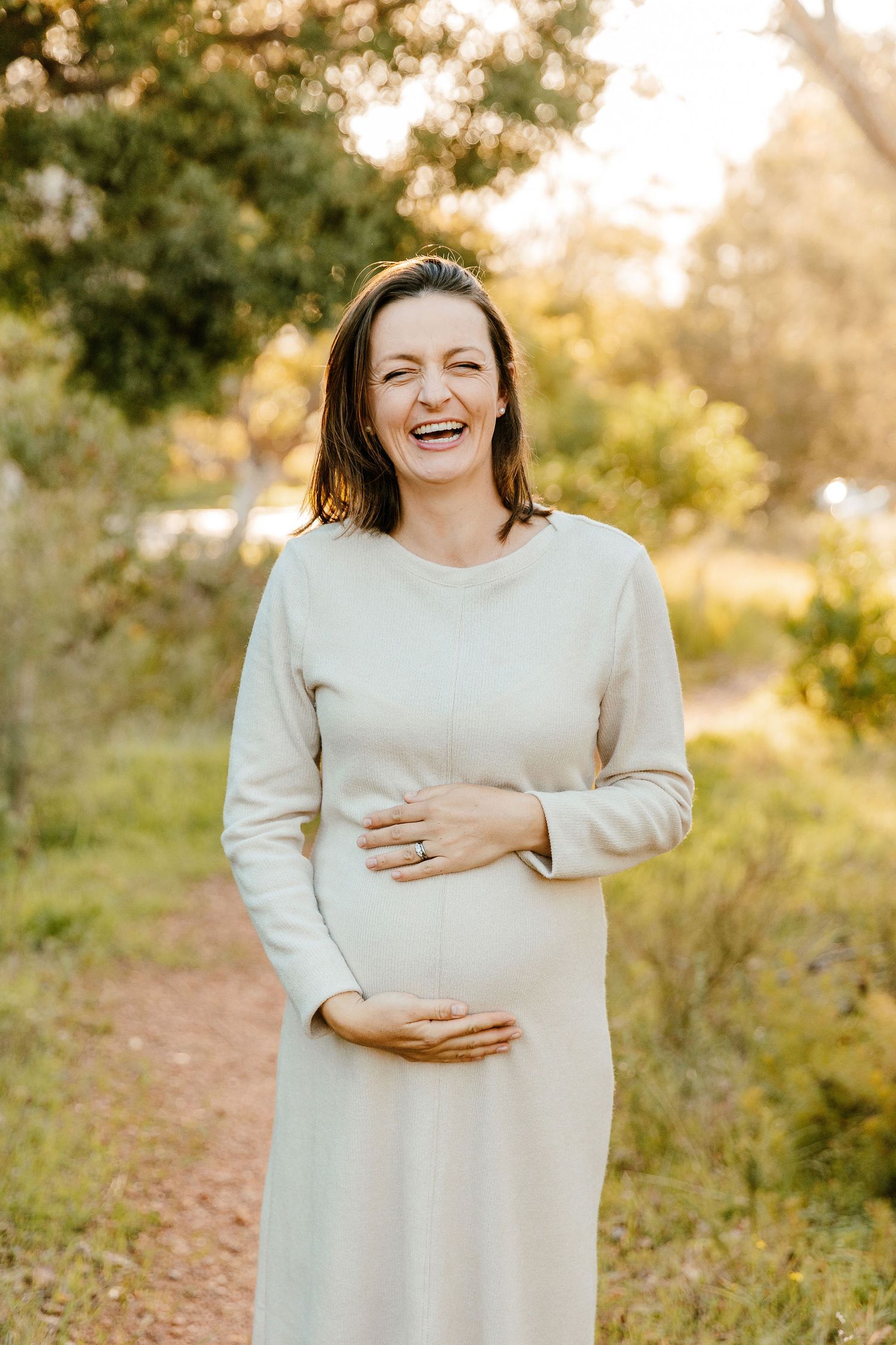 Laughing pregnant woman in white dress cradling baby bump outdoors in golden sunlight.