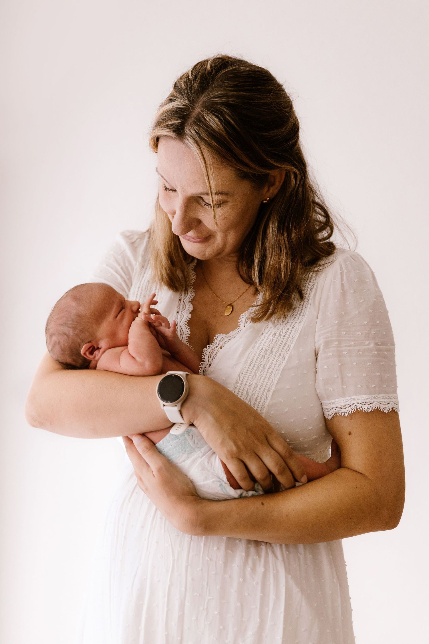 A smiling mother in a white lace dress tenderly holds her newborn baby against a soft white background.