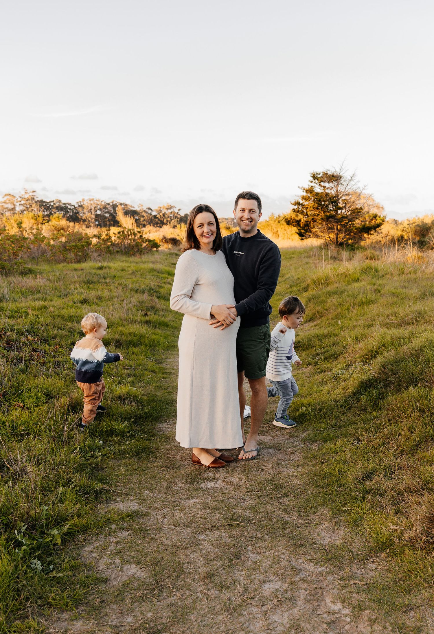 Pregnant couple poses outdoors on a grassy path at golden hour with two young children playing nearby.