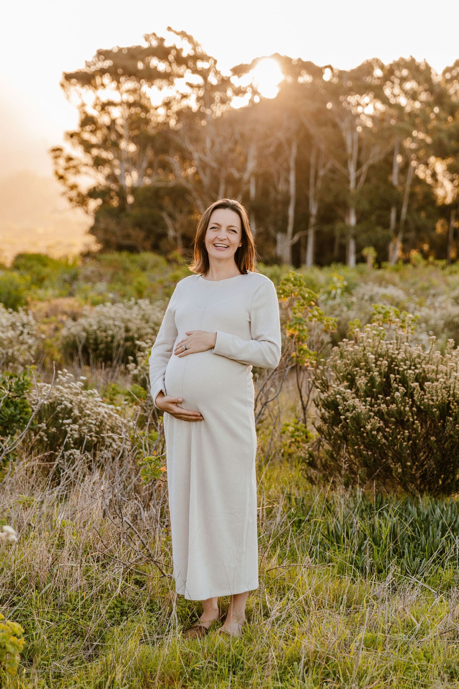 Smiling pregnant woman in white dress cradling baby bump outdoors at golden hour with trees in background.