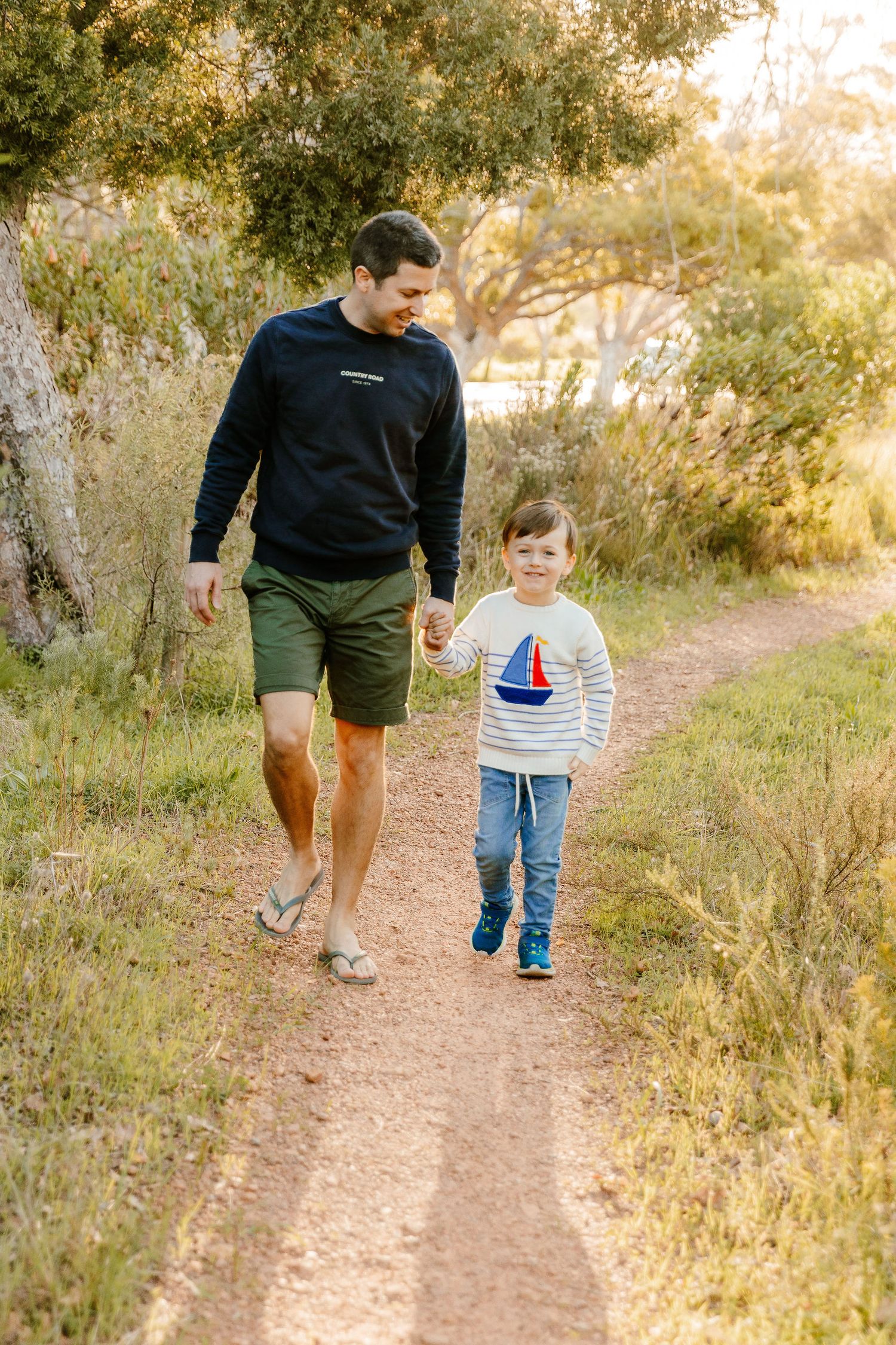 Father in a black sweatshirt and green shorts walking hand in hand with his young son on a trail.
