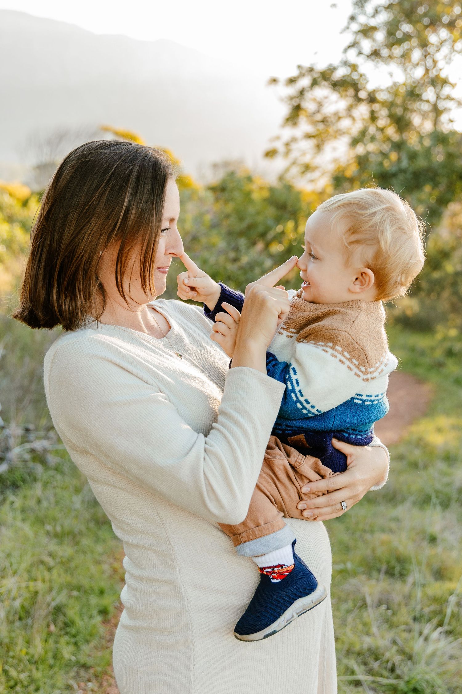 Mom and toddler touching noses playfully while being held in a sunny outdoor setting.