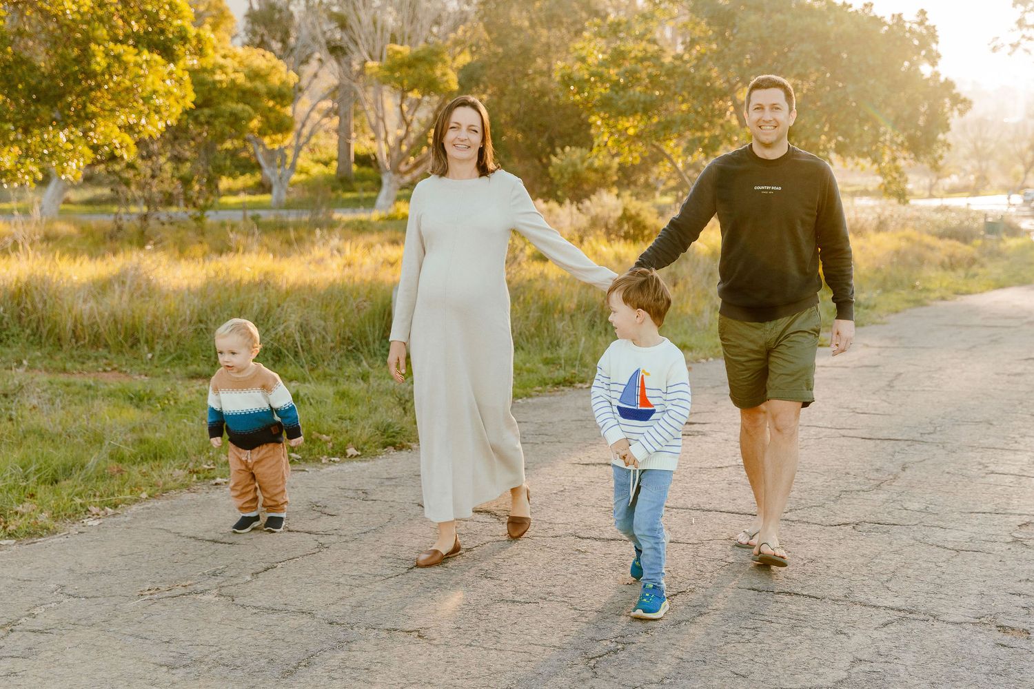 Pregnant woman and husband walking with two young sons on a sun-drenched outdoor pathway.