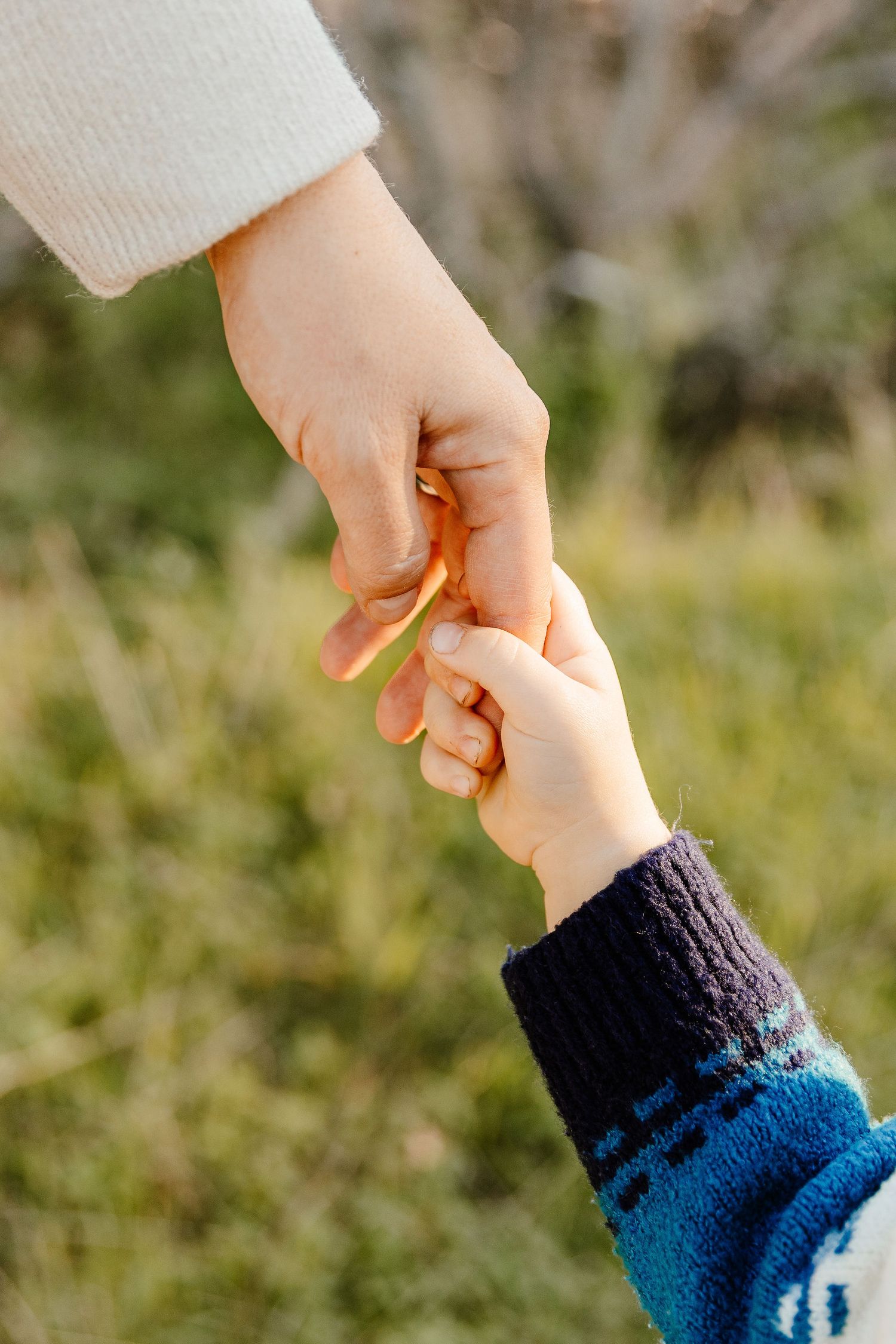 Close-up of adult and child holding hands in a grassy outdoor setting, wearing cozy knit sleeves.