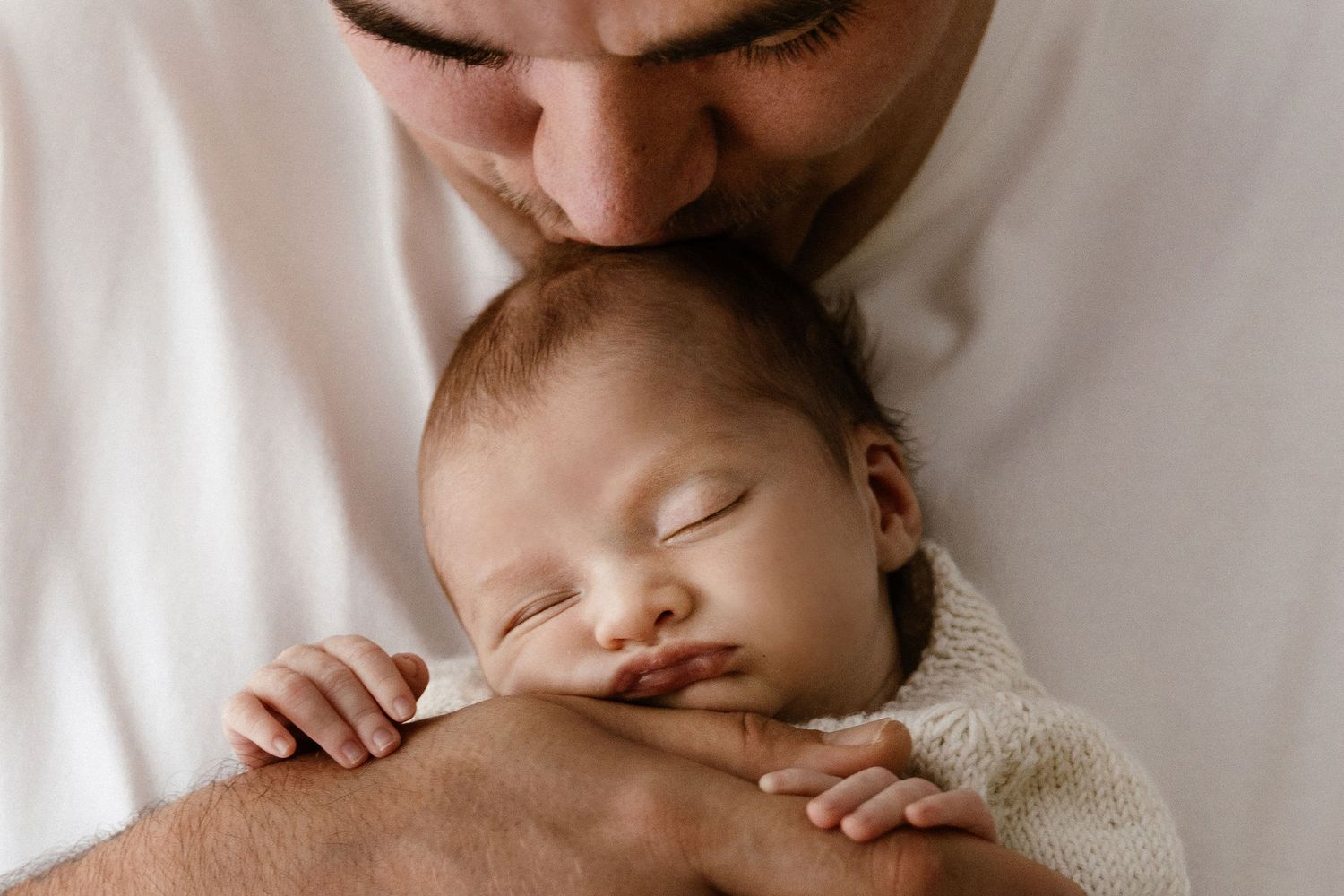 Father gently kissing sleeping newborn baby wrapped in soft white blanket.