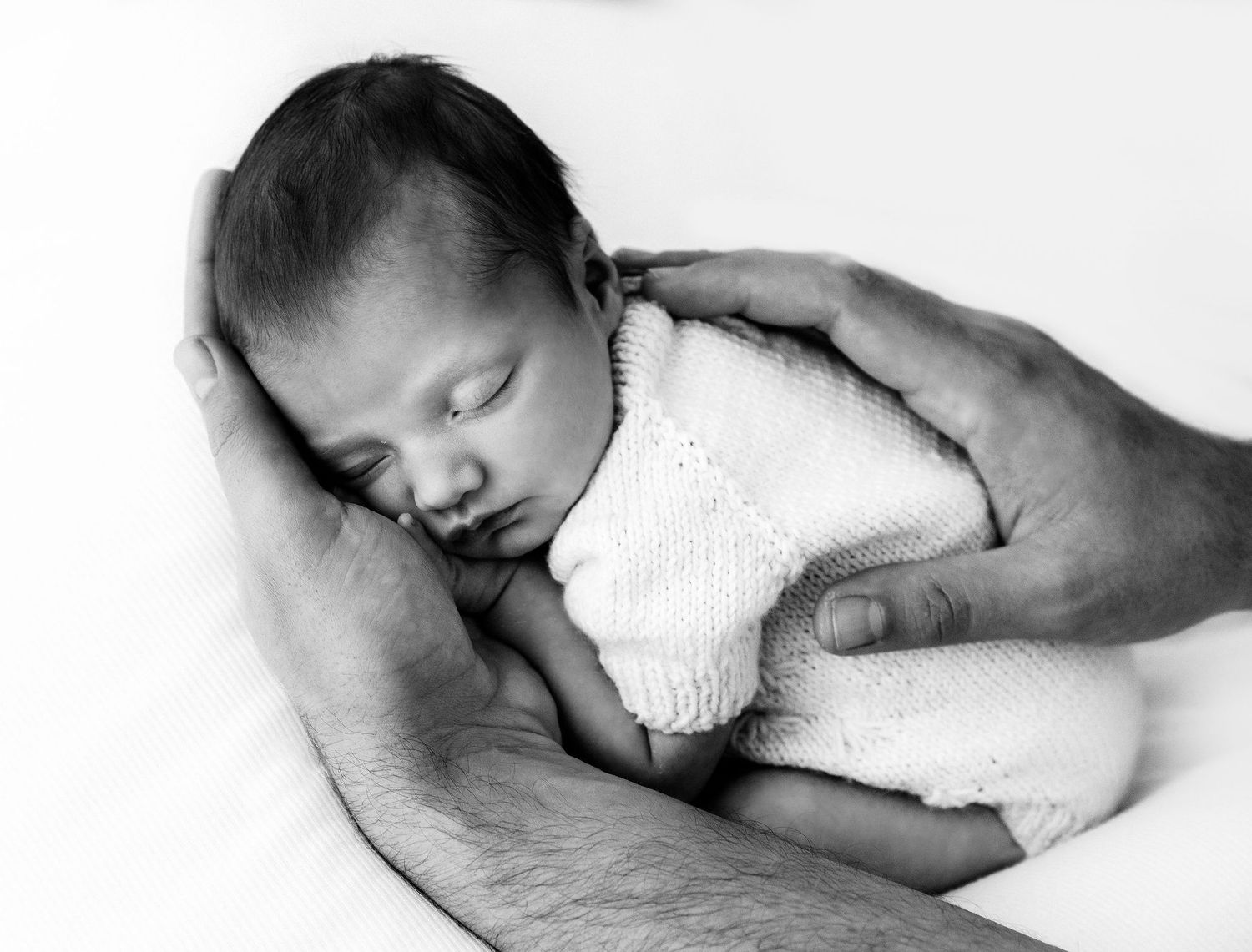 Black and white newborn photo of sleeping baby in knit outfit cradled gently in fathers hands.