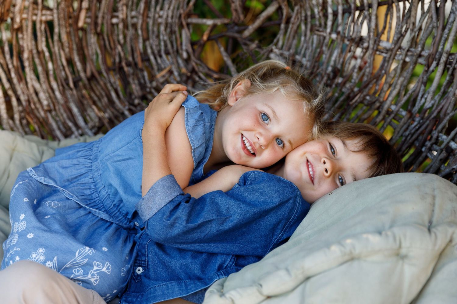Two smiling children in blue outfits cuddle together in a rustic woven nest, sharing a joyful moment outdoors.