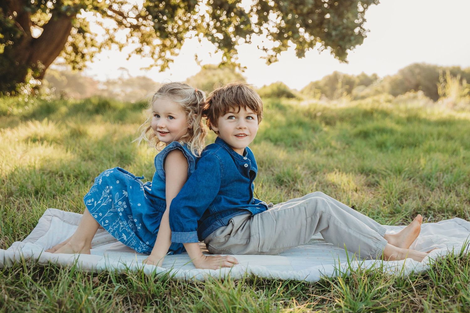 Young brother and sister sitting back-to-back on a blanket in a sunny green meadow.