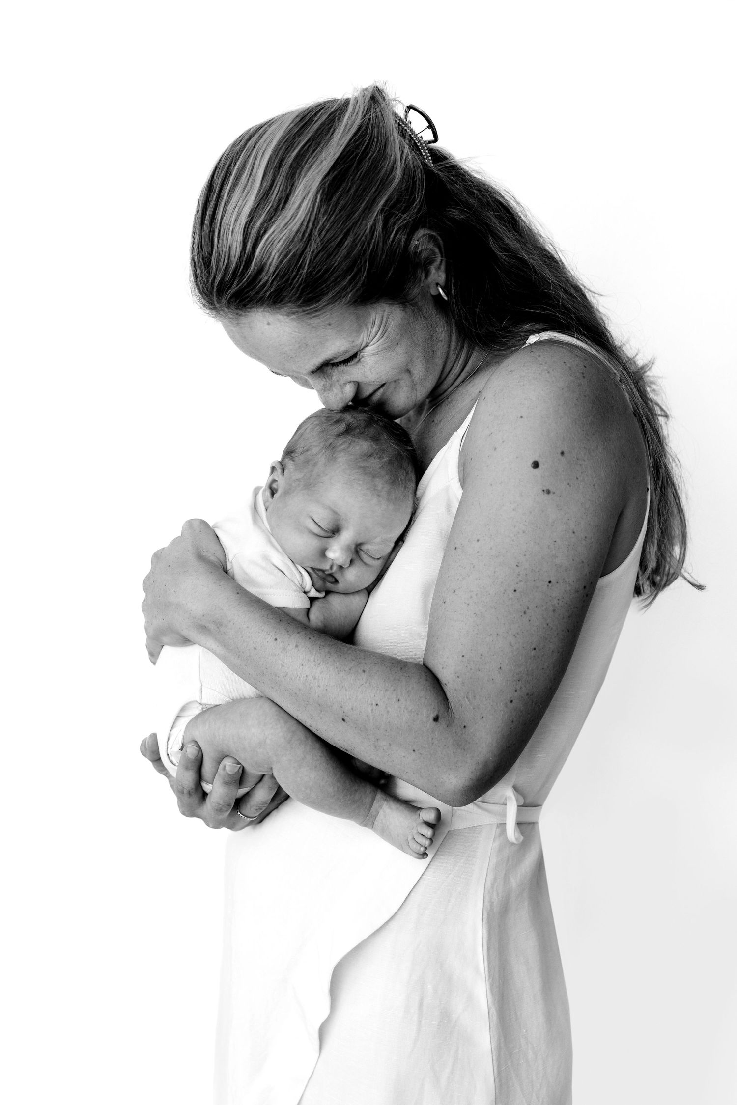Black and white photo of a mother tenderly holding and kissing her sleeping newborn baby wrapped in white cloth.