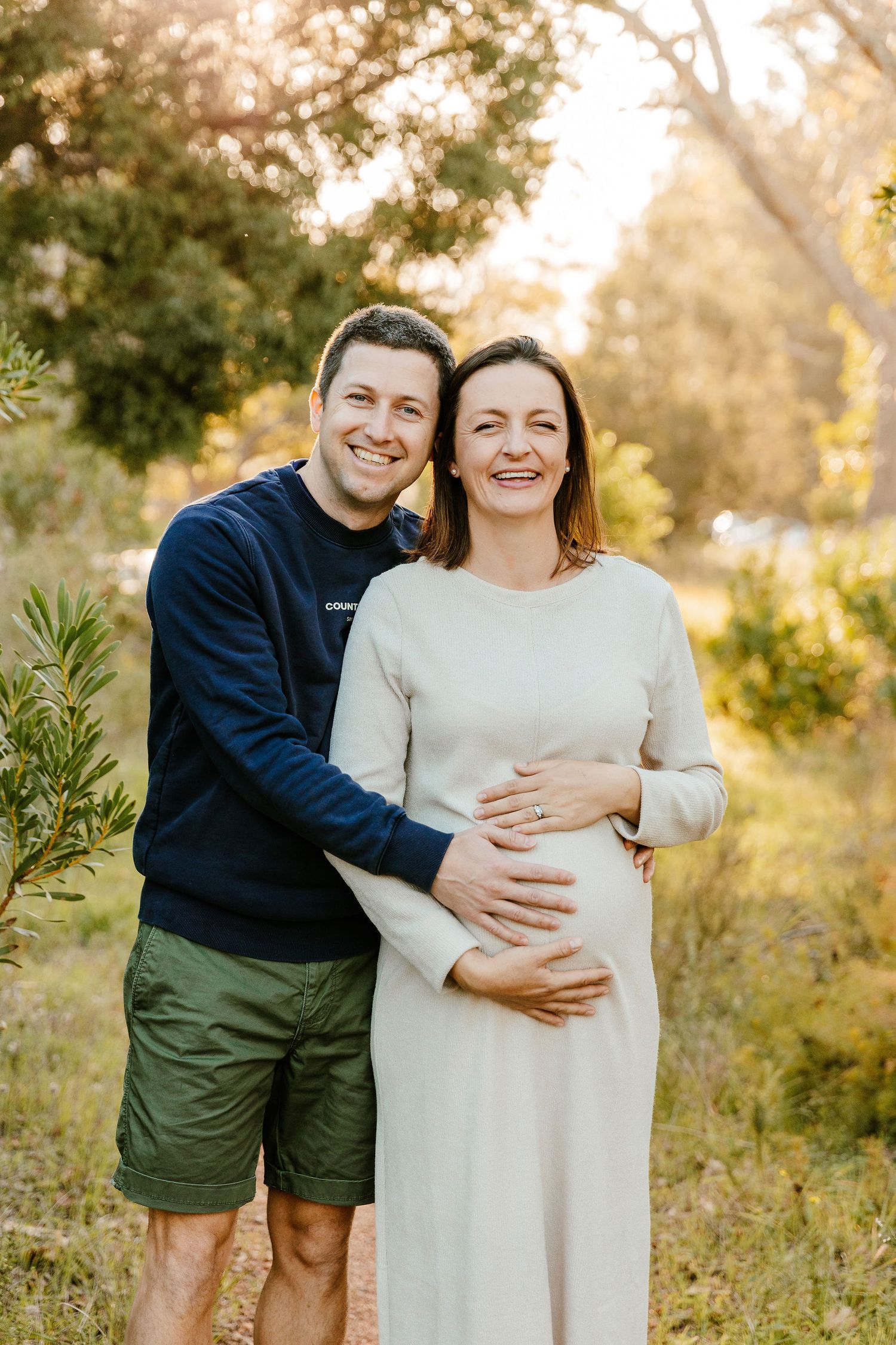 Smiling couple outdoors, pregnant woman in cream dress cradling baby bump, man in navy sweater embracing her in golden sunlight.