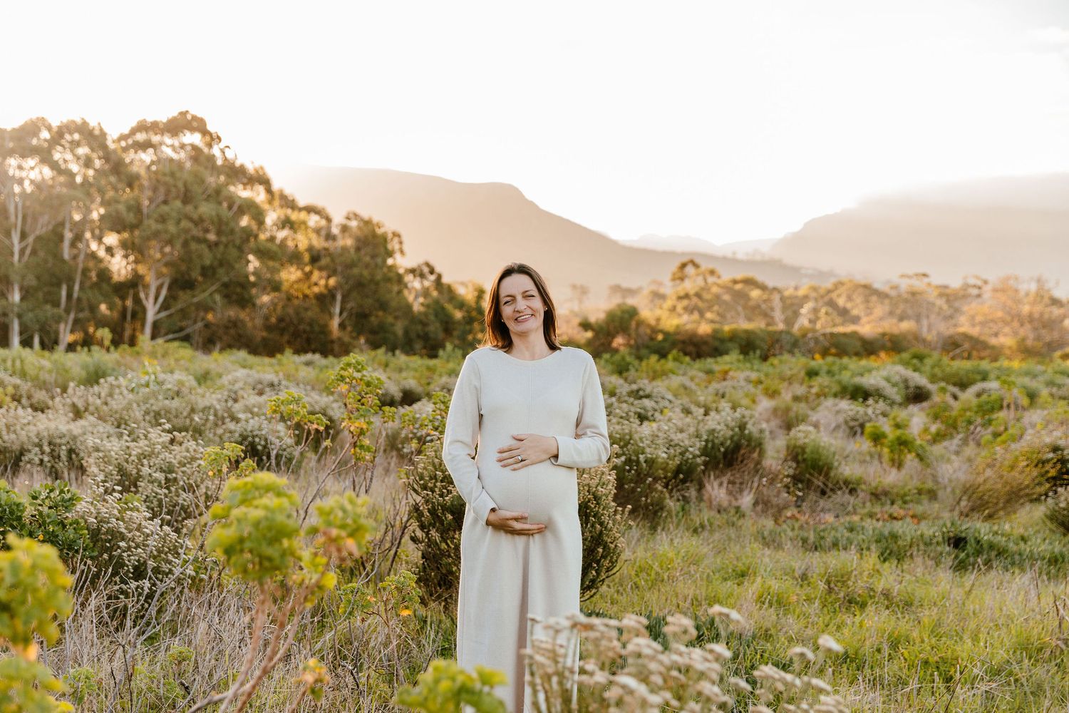 Pregnant woman in white dress smiling outdoors in golden sunlight with mountains and greenery in background.