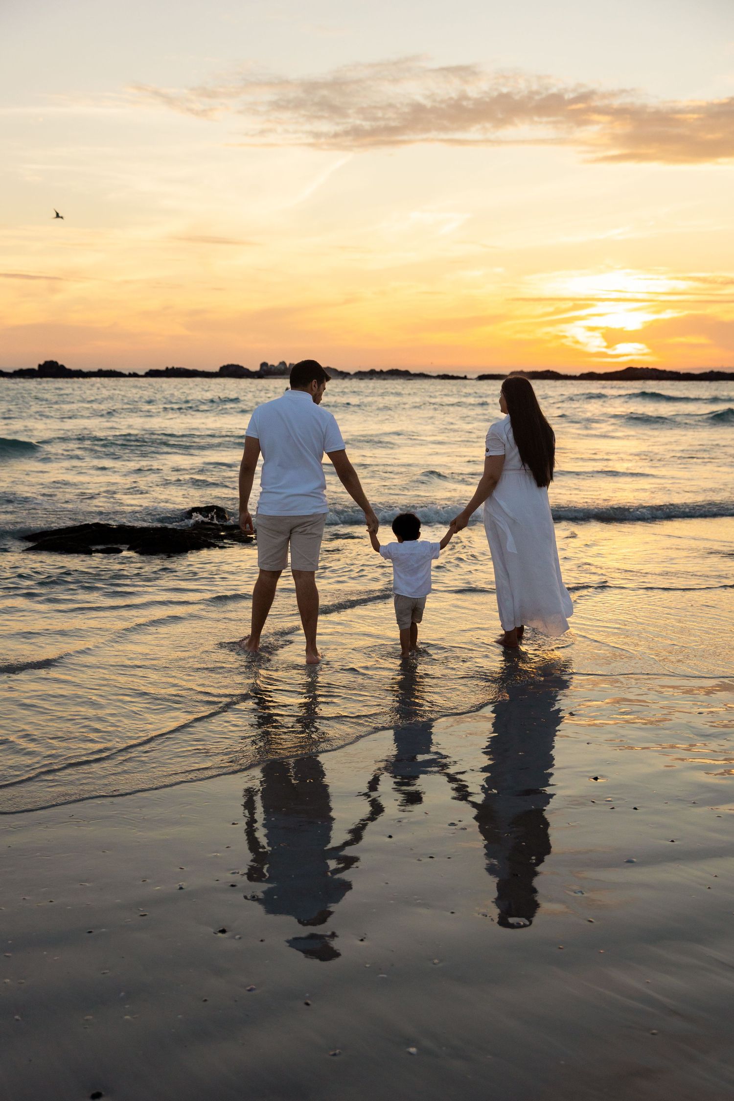 Family of three holding hands on beach at sunset, parents and toddler in white outfits with golden ocean reflection.