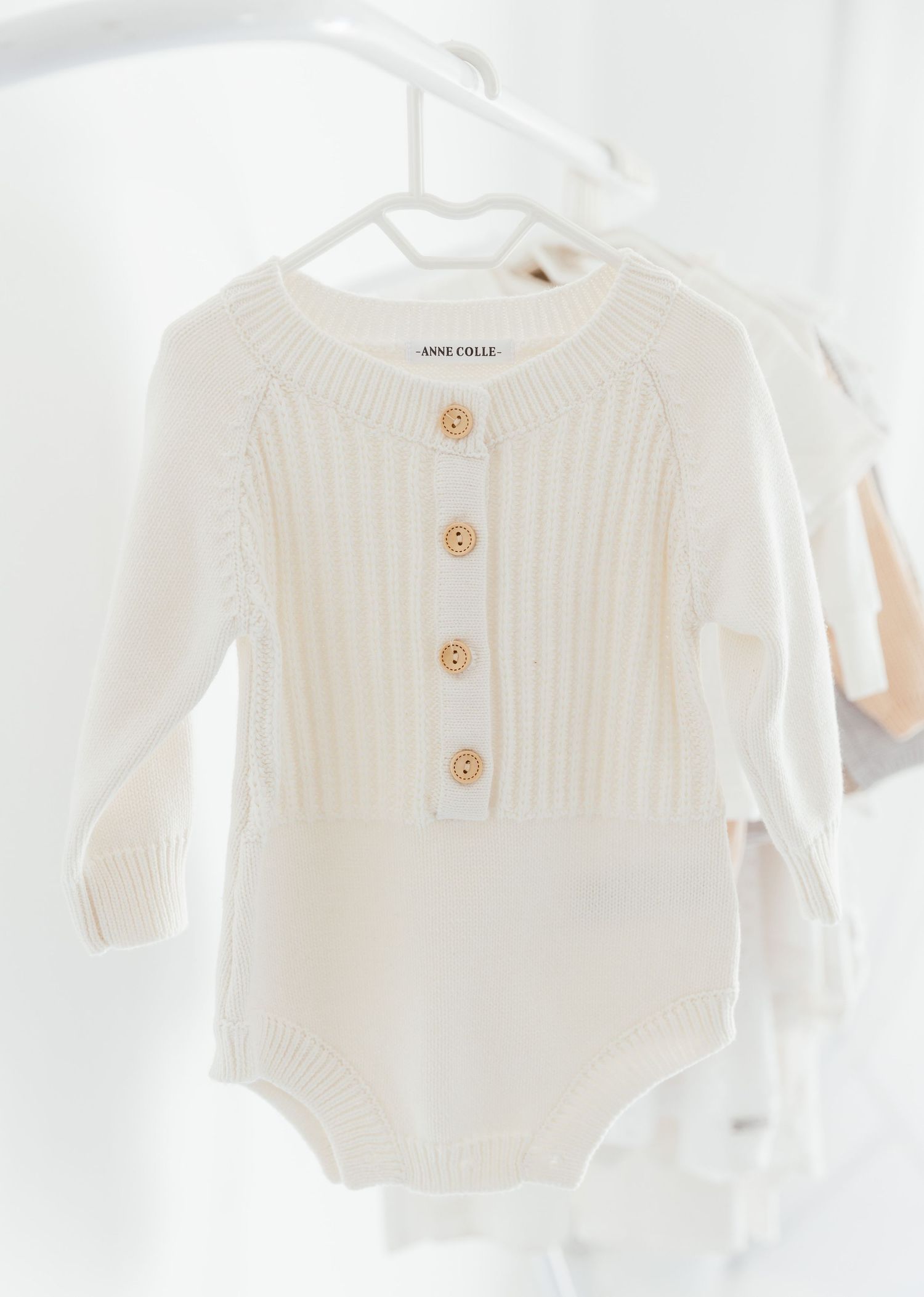 Cream knit baby romper with wooden buttons and long sleeves by Anne Colle, hanging on a white hanger.