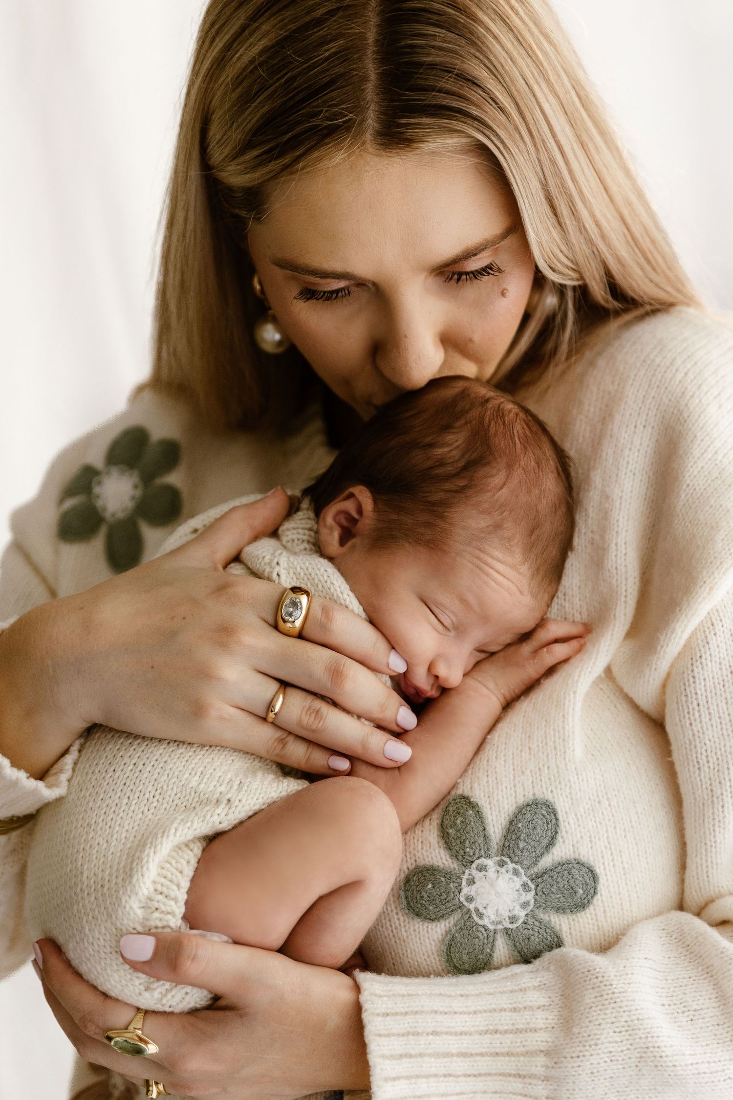 Mother in floral cream sweater gently kissing her sleeping newborn baby wrapped in a knit blanket.
