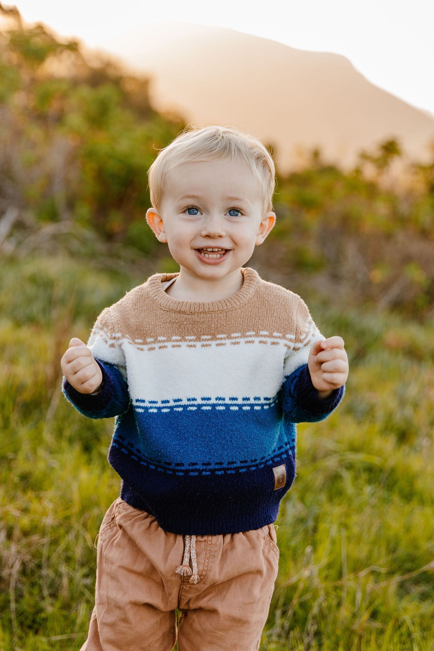 Happy blonde toddler boy smiling in a blue and tan striped knit sweater in a sunny green field.
