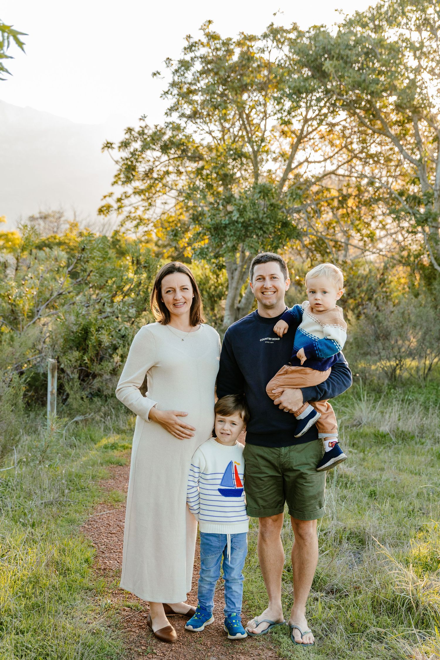 Happy family of four posing outdoors at sunset, pregnant mother in white dress.