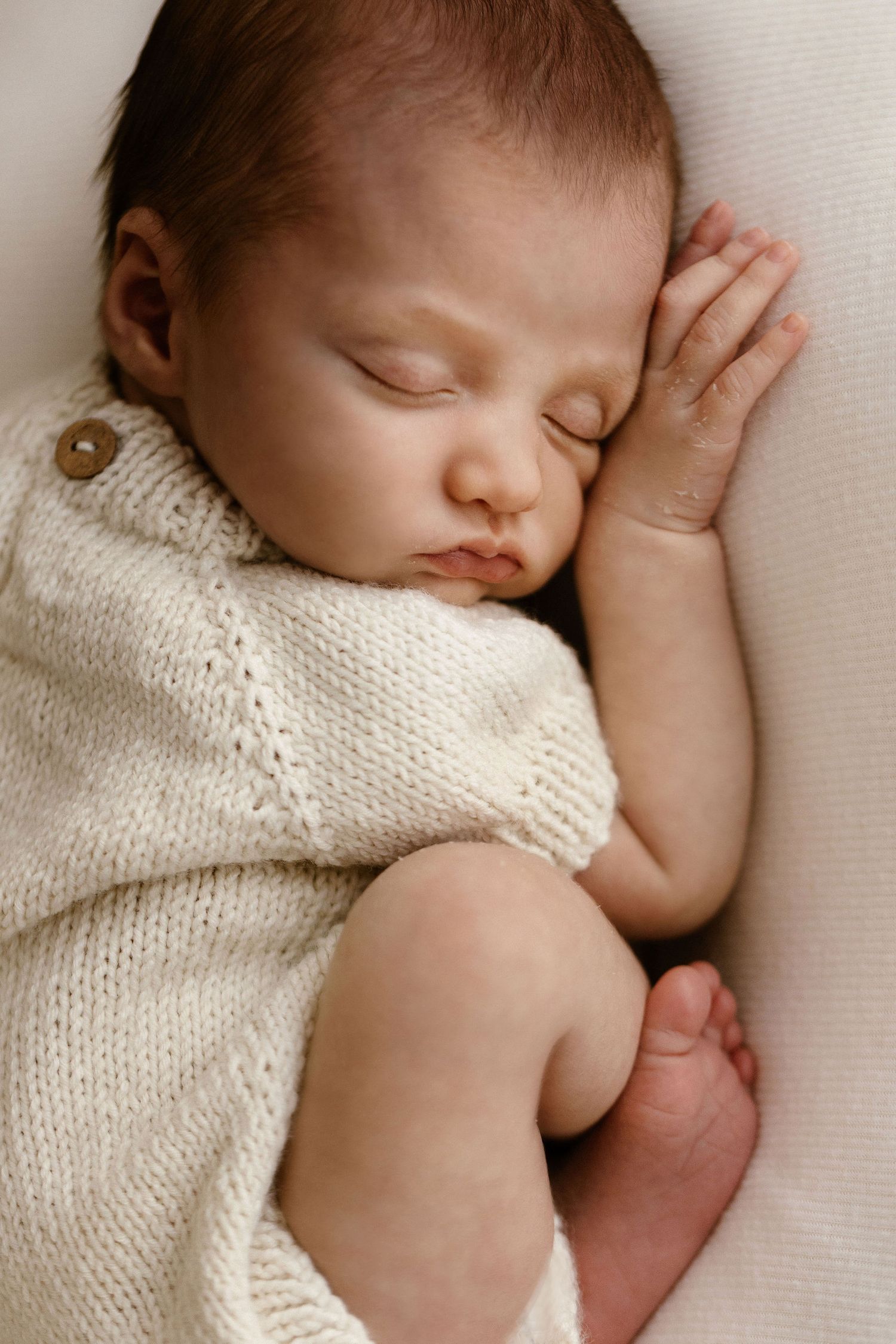 Serene newborn baby sleeping in cream knit romper with hand near face.