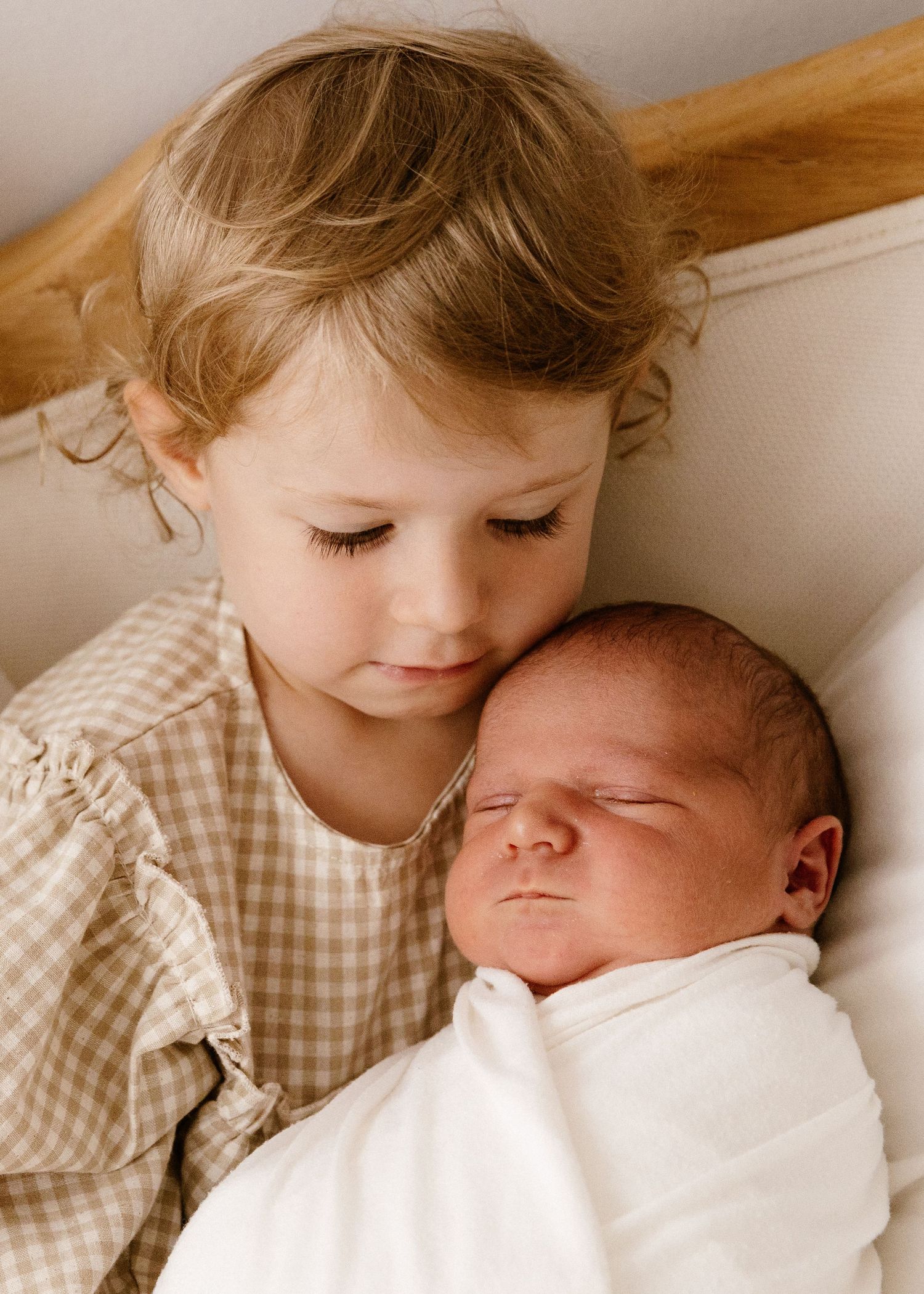 A young toddler girl tenderly holds a sleeping newborn baby swaddled in white.