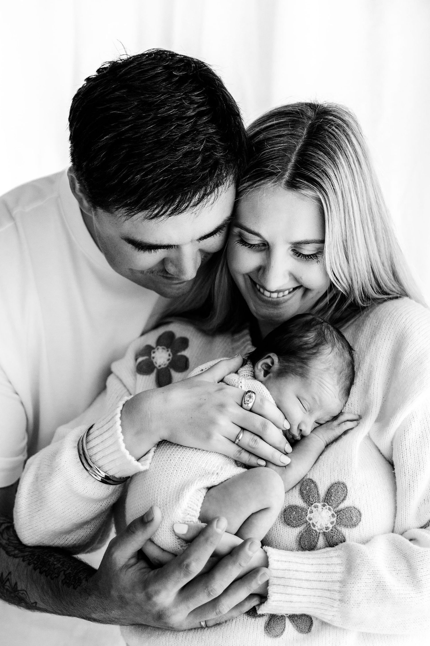 Black and white photo of smiling parents tenderly holding their sleeping newborn baby close together.