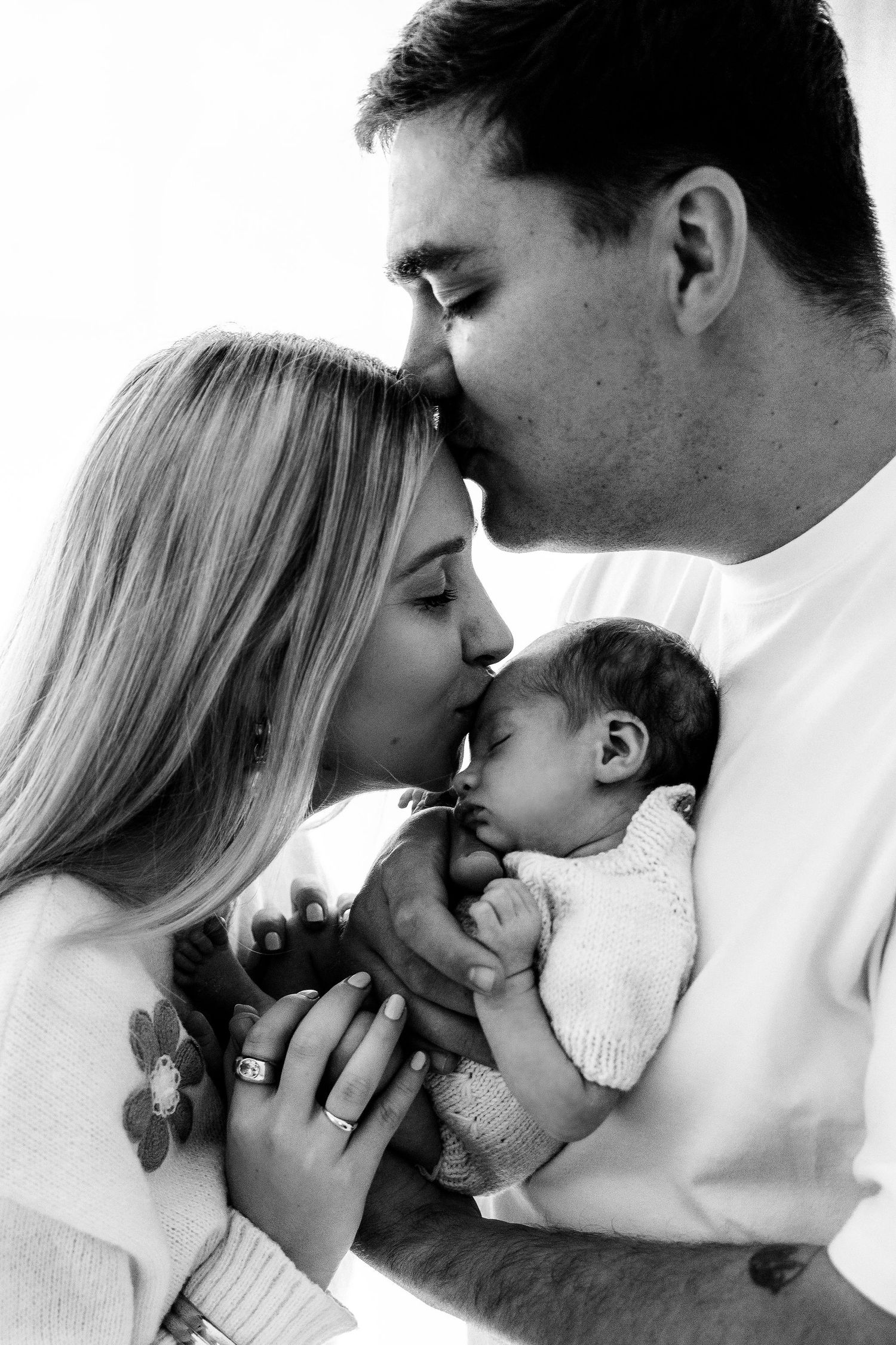 Black and white photo of parents kissing their newborn baby, a tender family portrait moment.