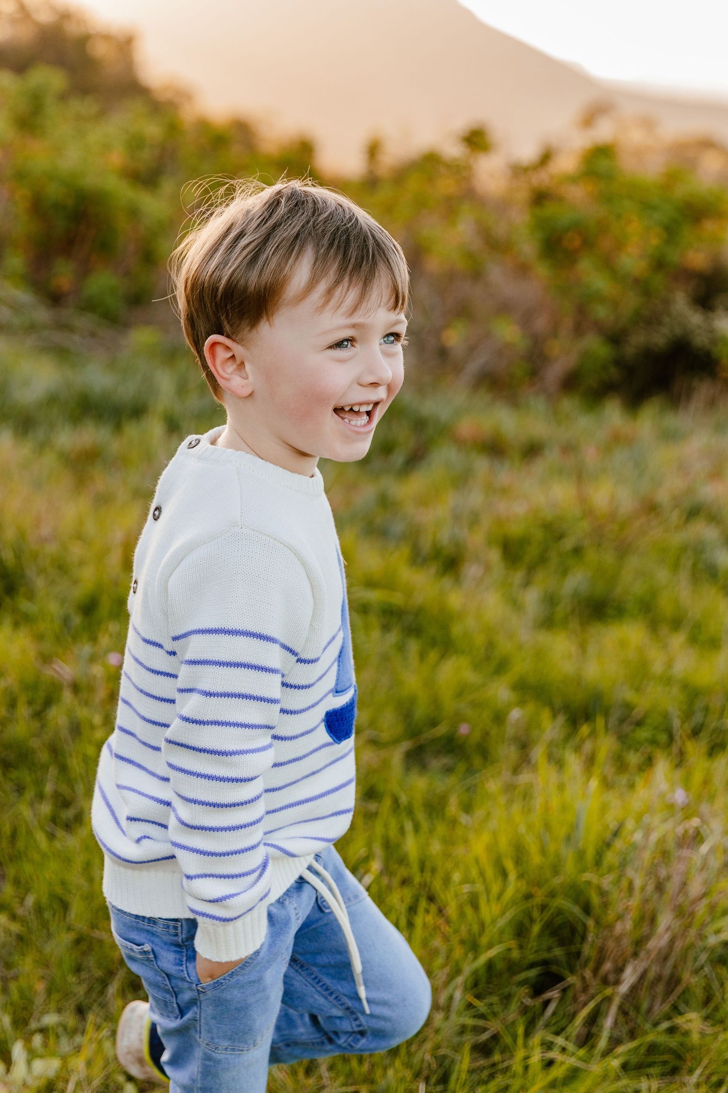 Smiling young boy in white striped sweater and jeans playing in a sunny green field.