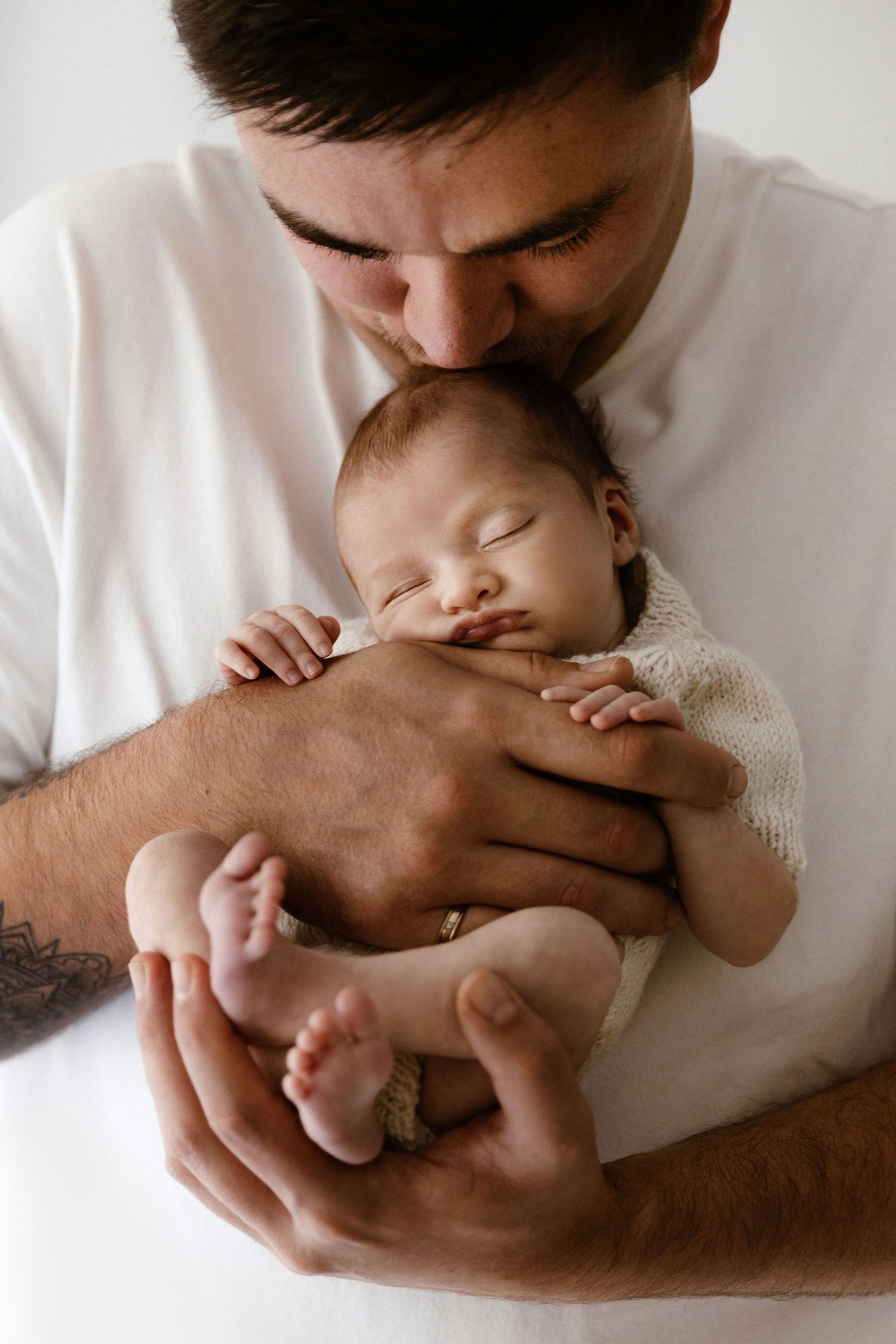 Loving father in white t-shirt kissing sleeping newborn baby held close to his chest.