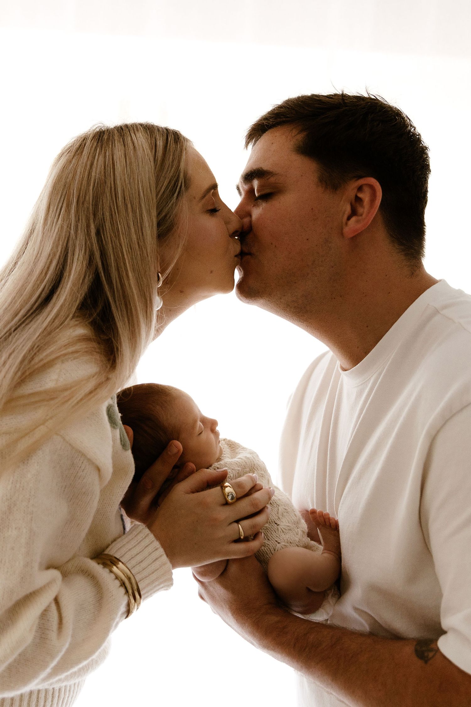Parents kiss while holding their newborn baby between them in bright light.