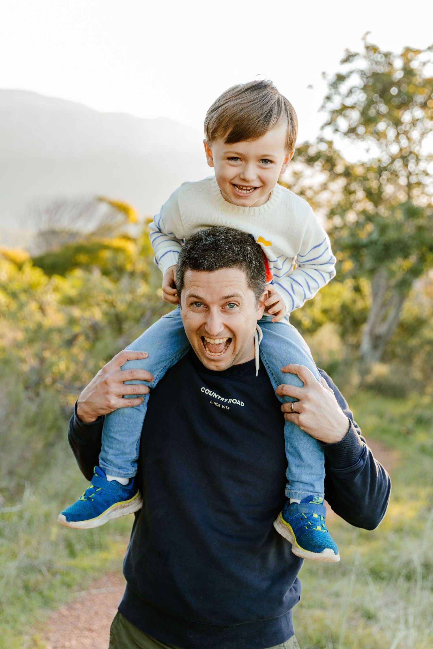 Laughing father with son sitting on his shoulders on a sunny outdoor nature trail.