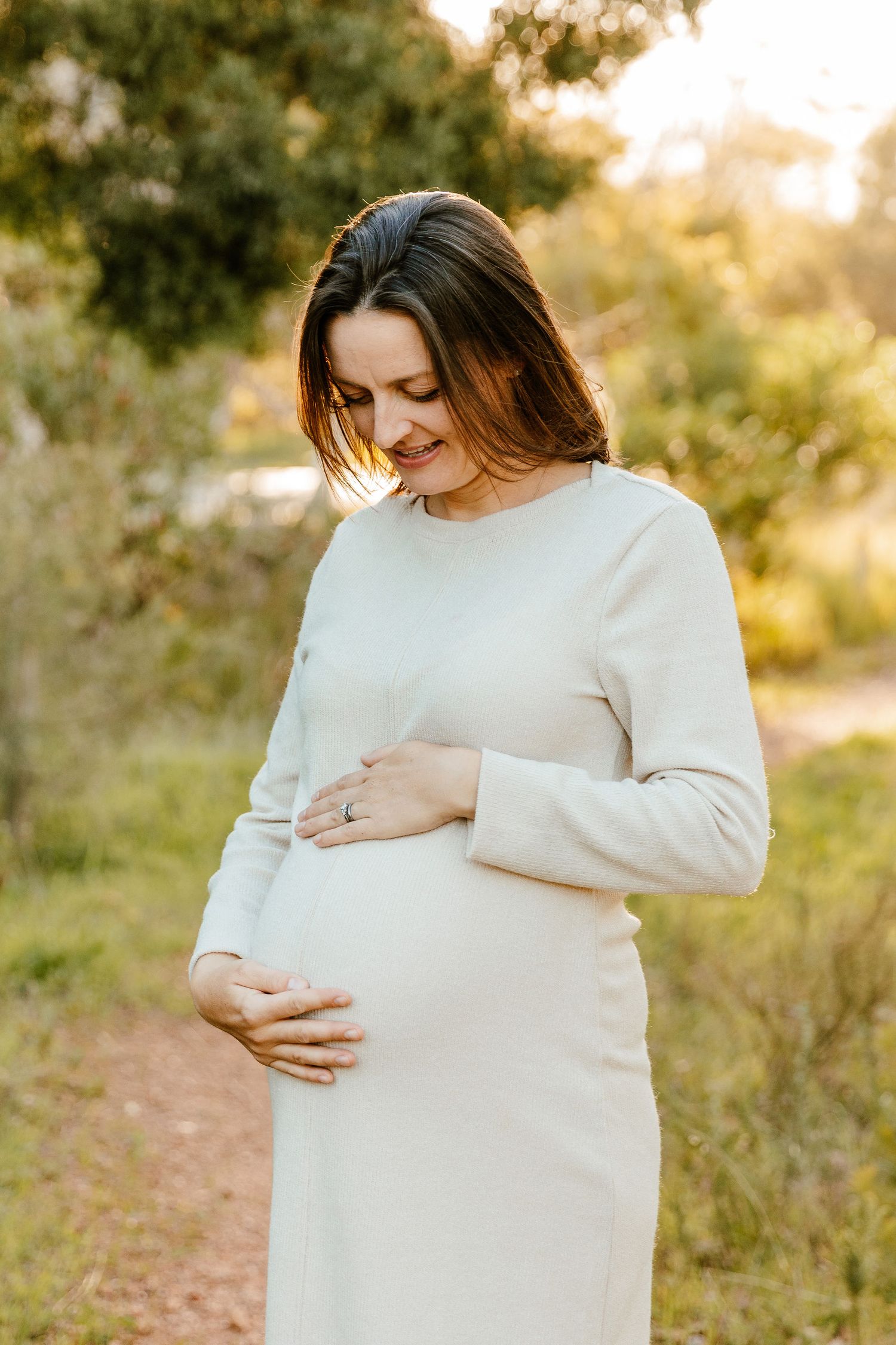 Pregnant woman in white dress cradling her baby bump outdoors in golden hour light.