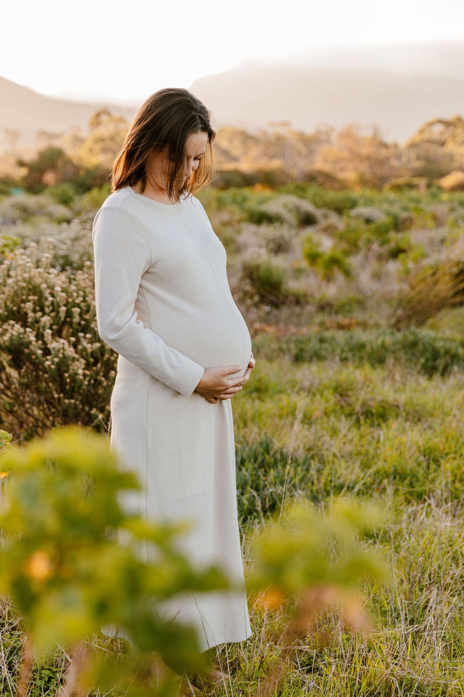 Pregnant woman in white dress cradling baby bump outdoors at golden hour with mountains and greenery.