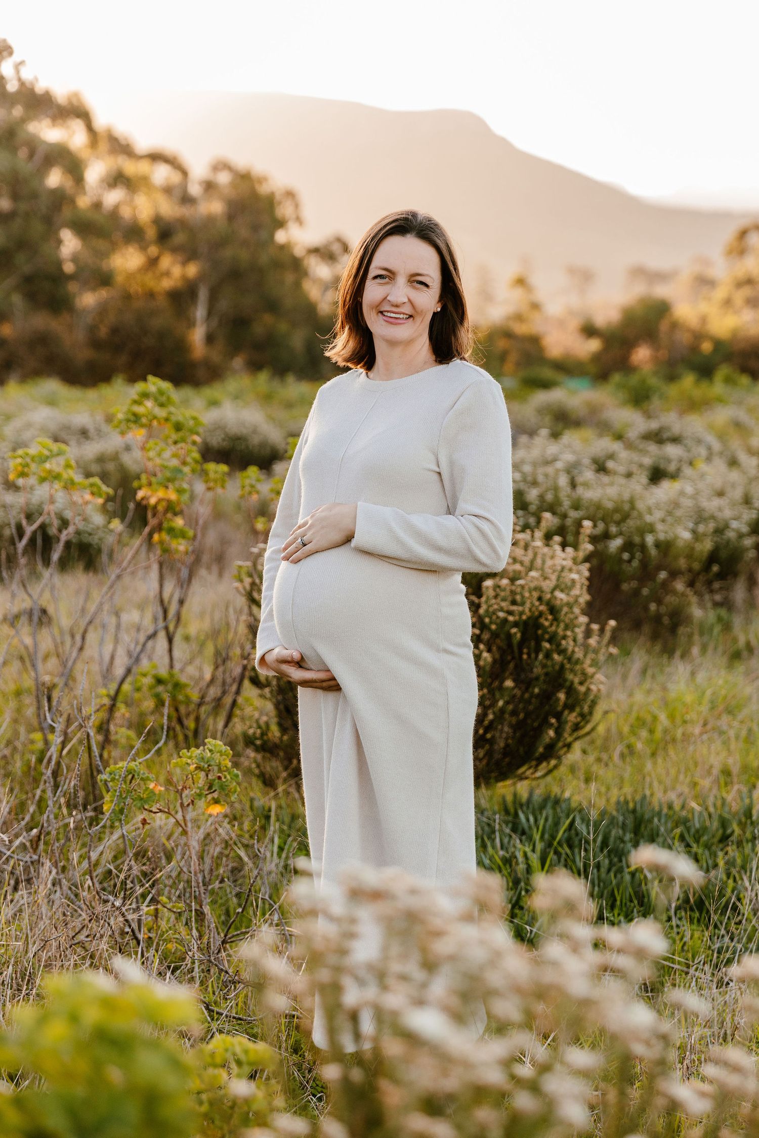 Smiling pregnant woman in white dress cradling baby bump outdoors at golden hour with mountains and greenery.