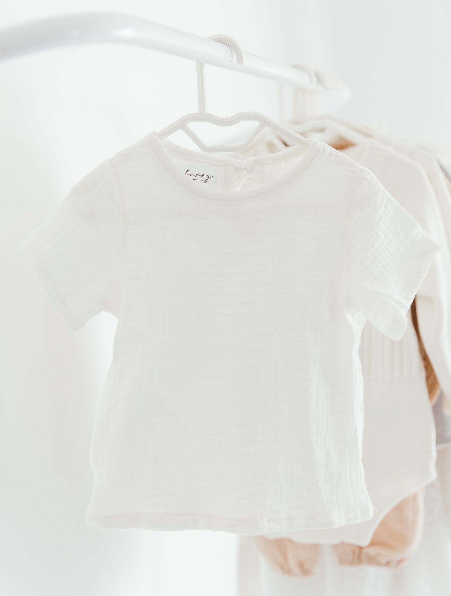 White short-sleeve baby t-shirt hanging on a hanger among neutral-toned children's clothing on a rack.