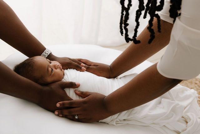 Two adults tenderly cradling a sleeping newborn baby wrapped in white on a soft white surface.