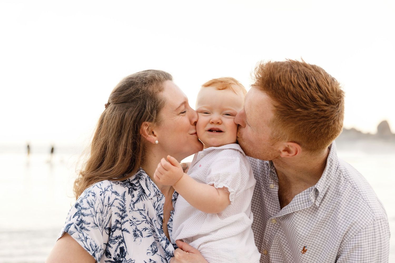 Happy redhead parents kissing their smiling baby on the cheeks at the beach.
