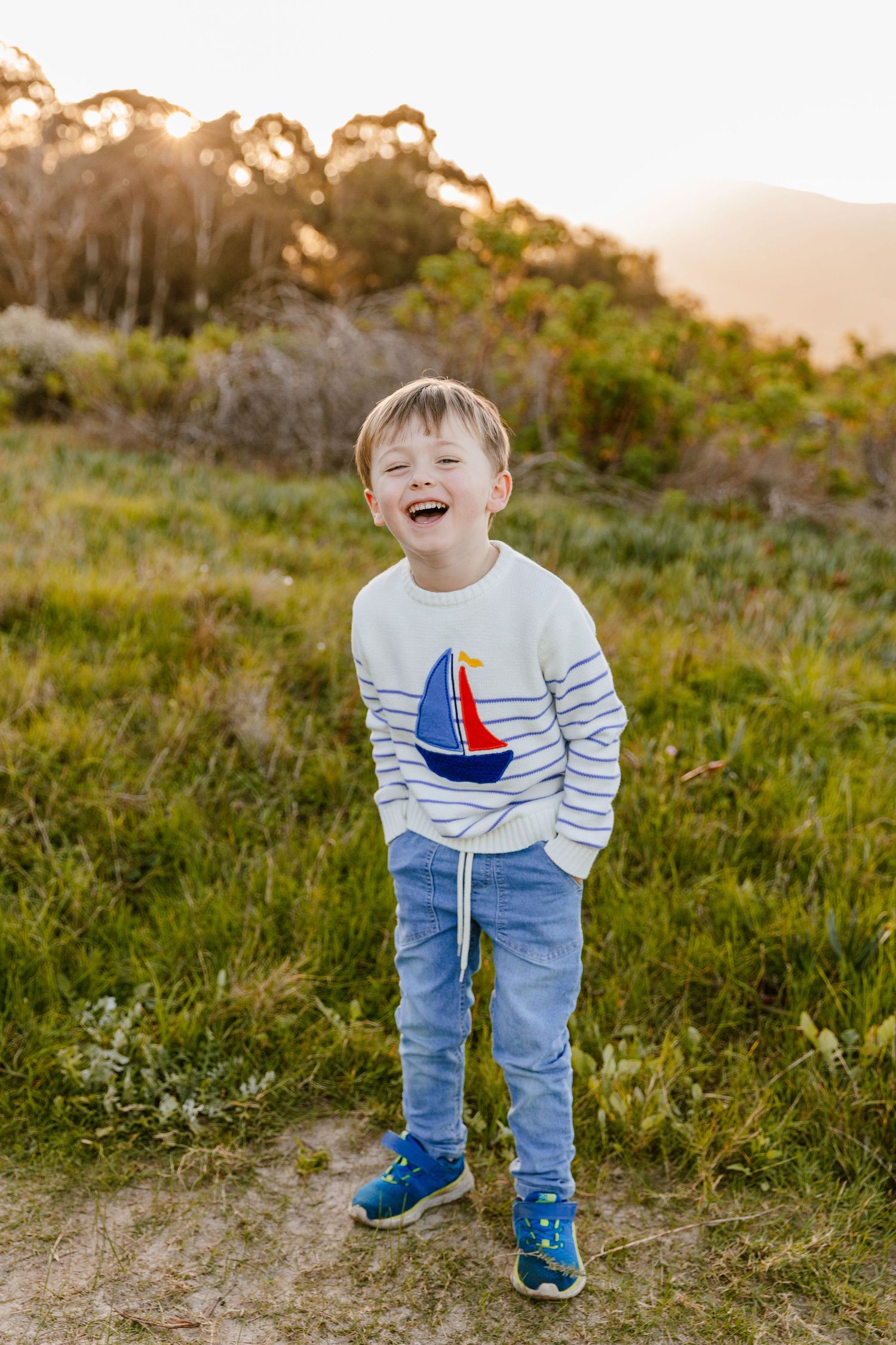 Joyful young boy in a sailboat sweater laughing outdoors in warm sunset light.