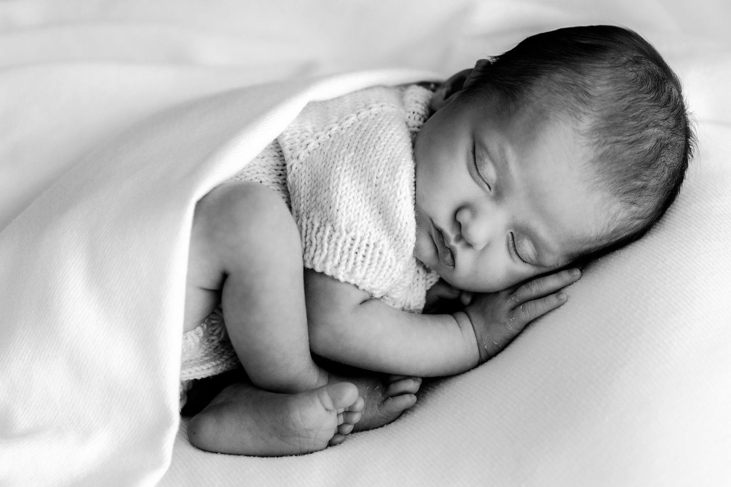 Black and white photo of a curled sleeping newborn baby wrapped snugly in a soft white blanket.