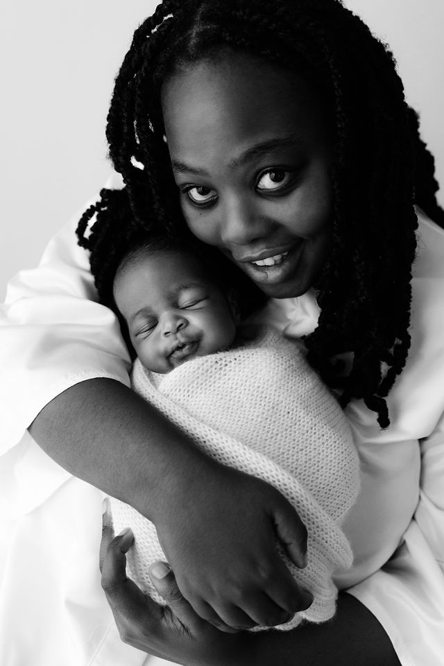 Black and white portrait of a smiling mother with braids holding her sleeping swaddled newborn baby close to her cheek.