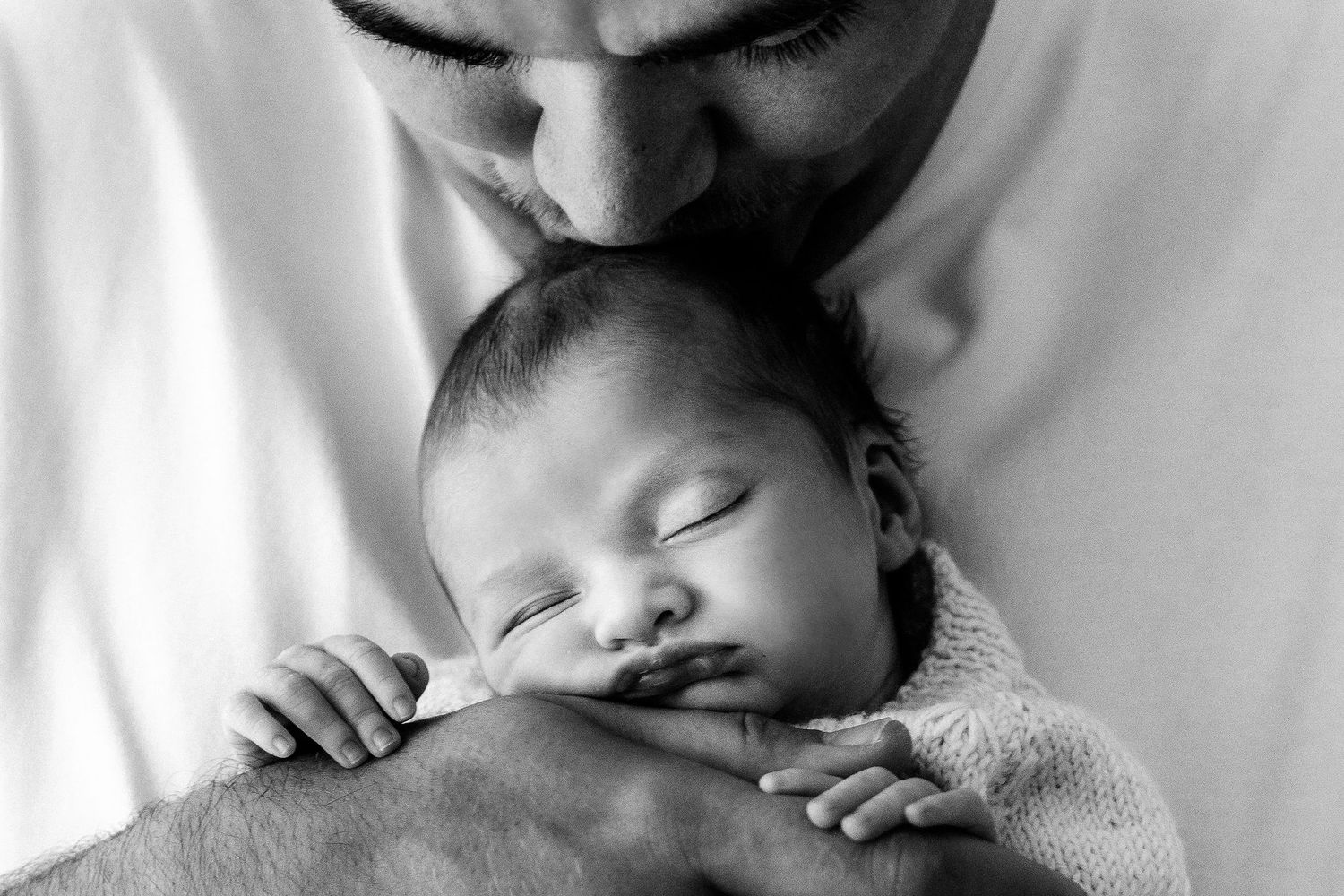 Black and white photo of a parent kissing a sleeping newborn baby wrapped in a knit blanket.
