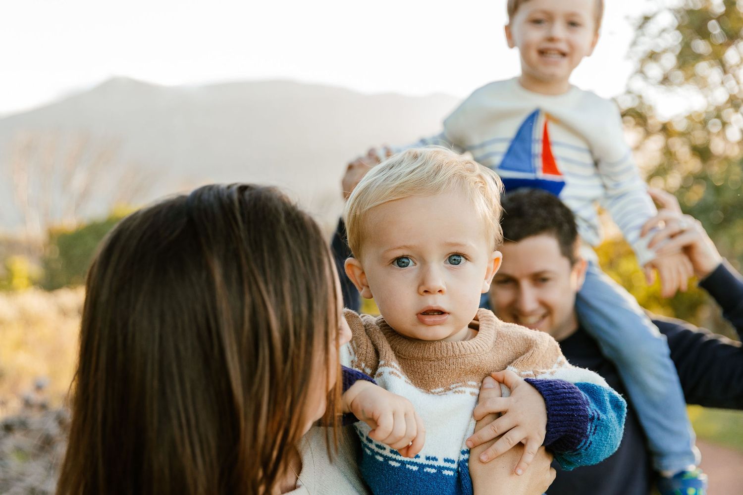 A mother kisses her blue-eyed toddler while father holds older child outdoors at golden hour.