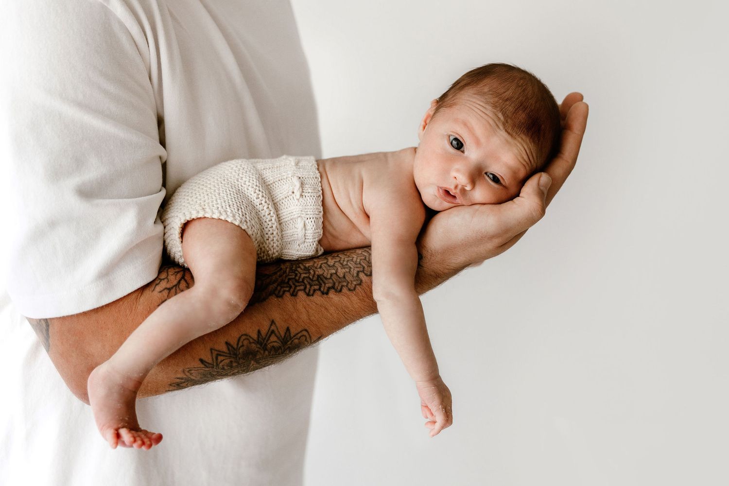 Tattooed father holding sleeping newborn baby on his forearm against white background.