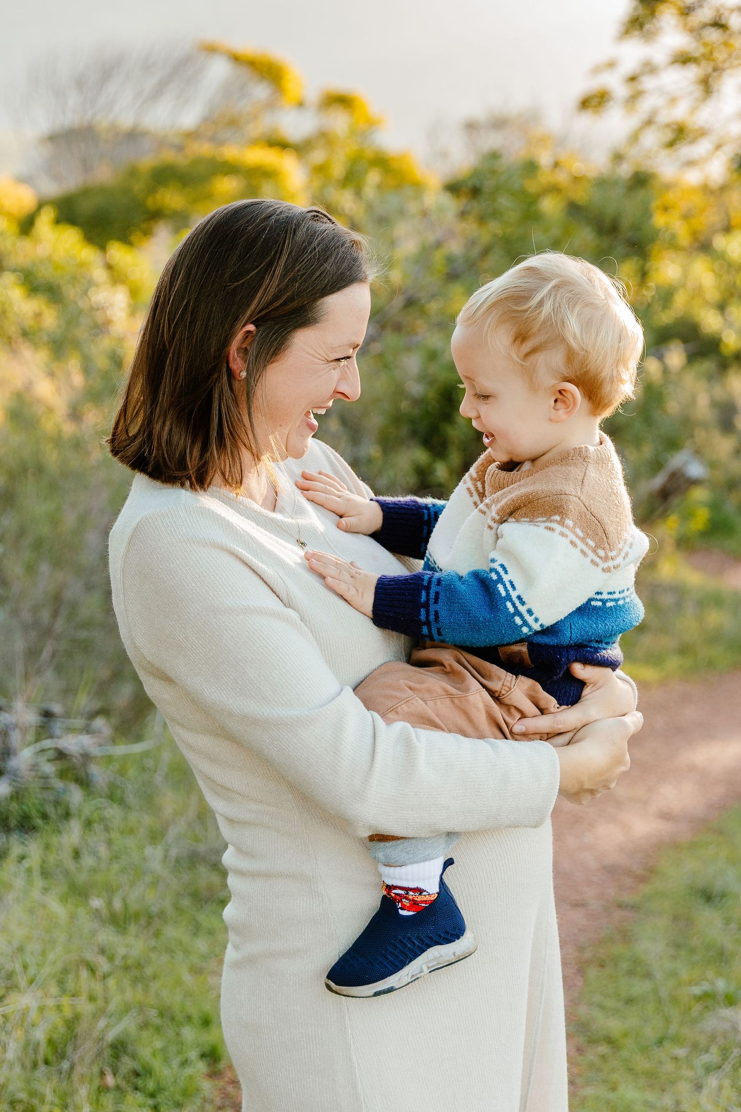 Laughing pregnant woman in white dress holds a happy blonde toddler outdoors at sunset.