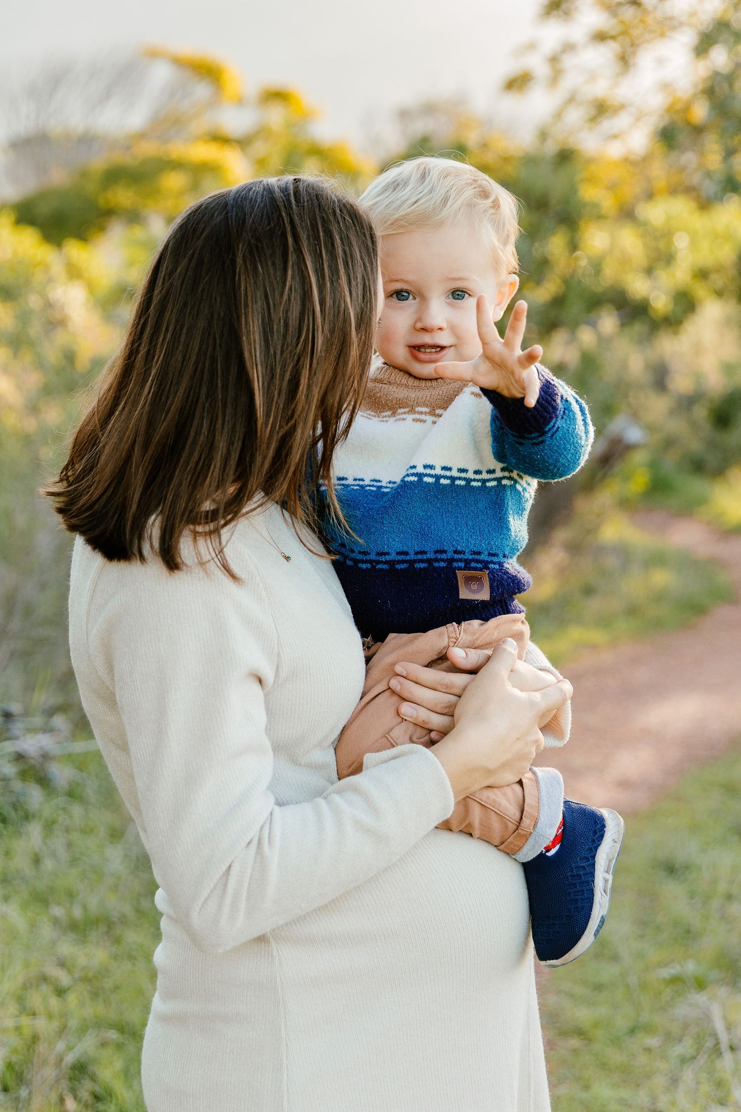 Pregnant woman holding a curious blonde toddler waving on a sunlit nature path.