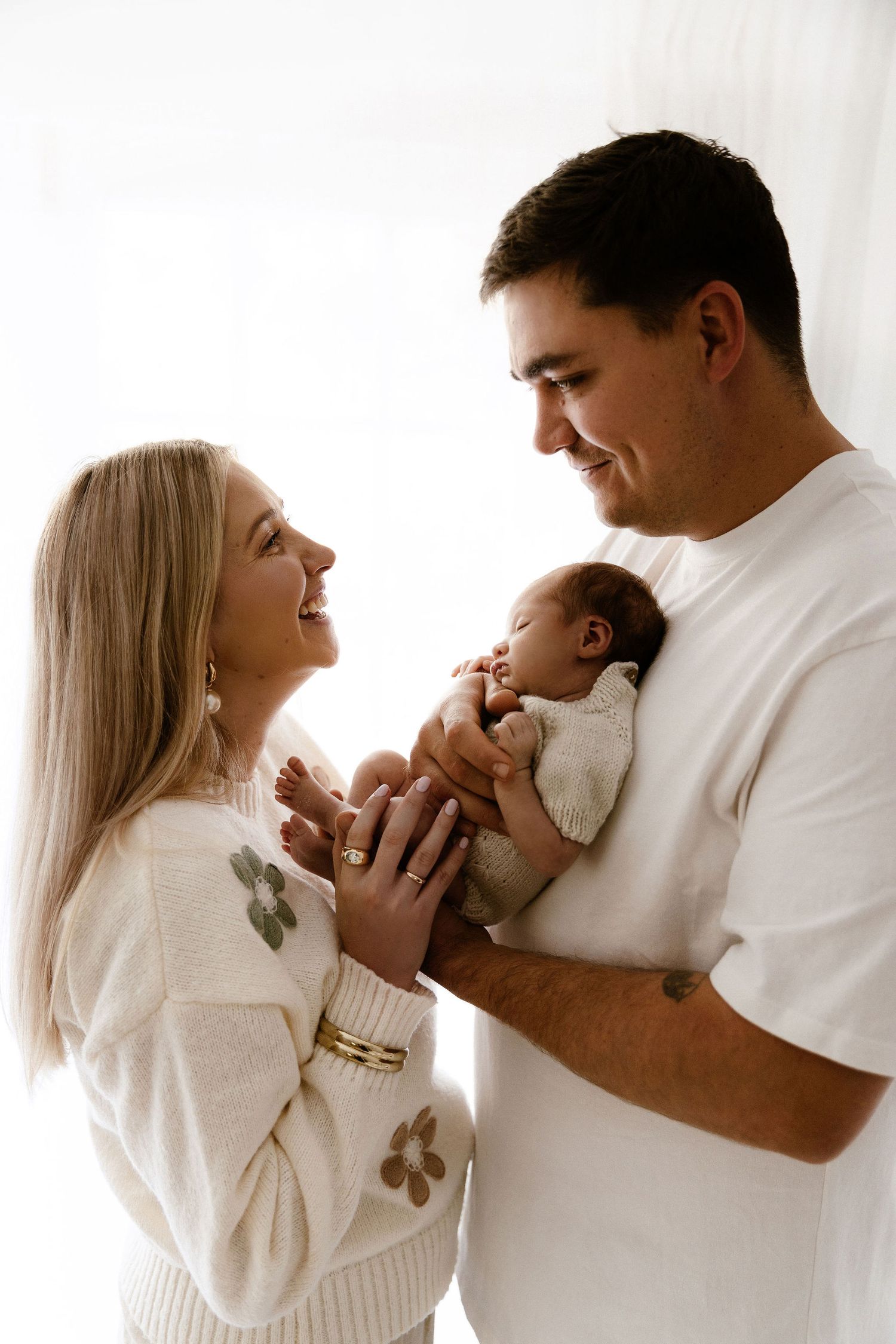 Smiling couple in white outfits holding their newborn baby together.
