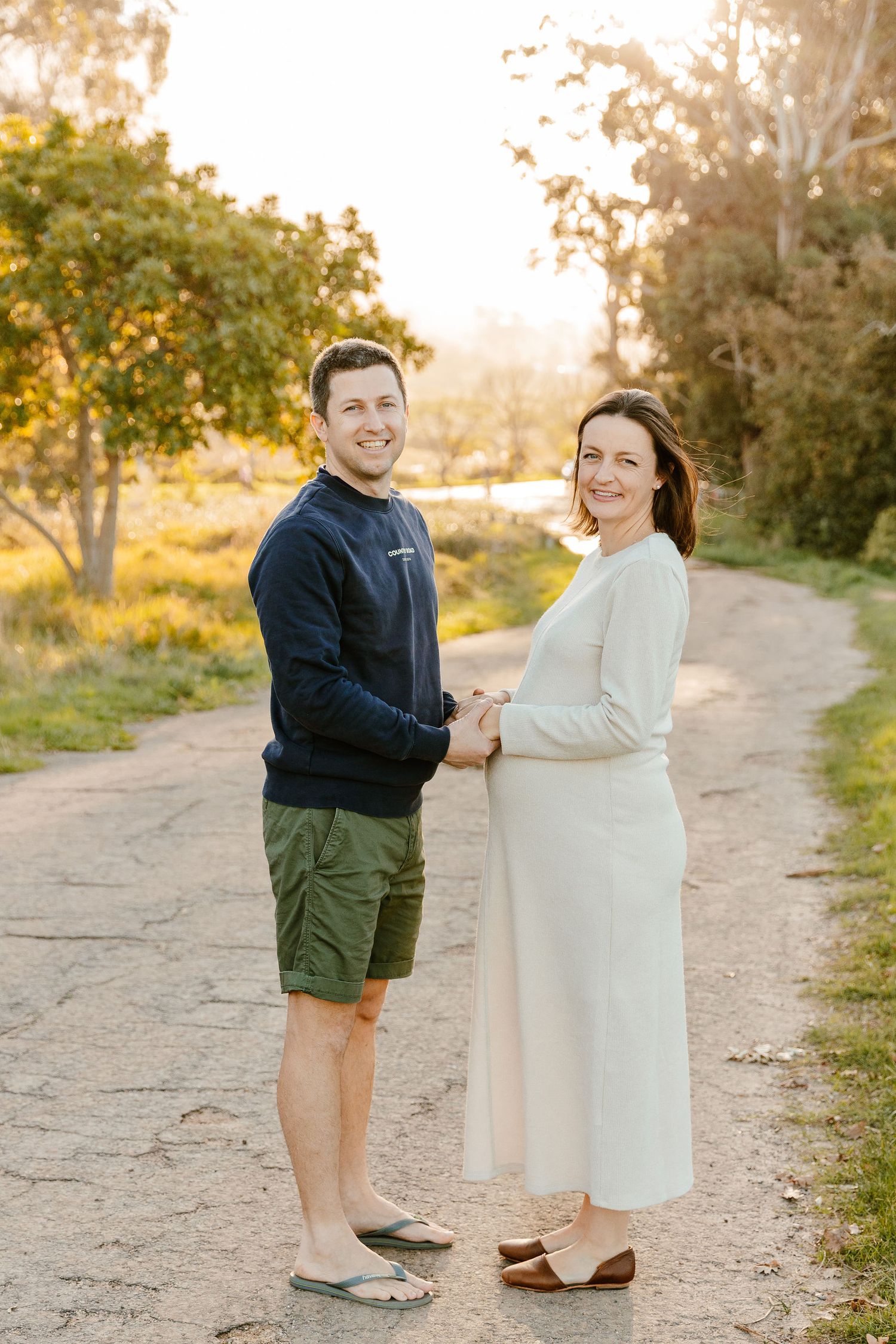 Smiling couple holding hands on a sunlit dirt path surrounded by golden trees during a maternity photo session.