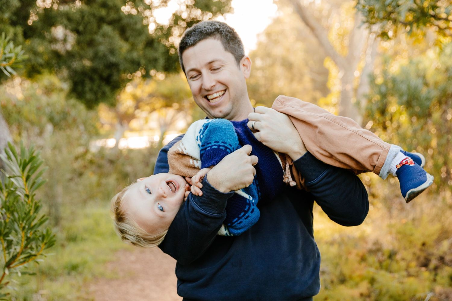 Laughing father playfully holding toddler upside down outdoors on a nature trail during golden hour.