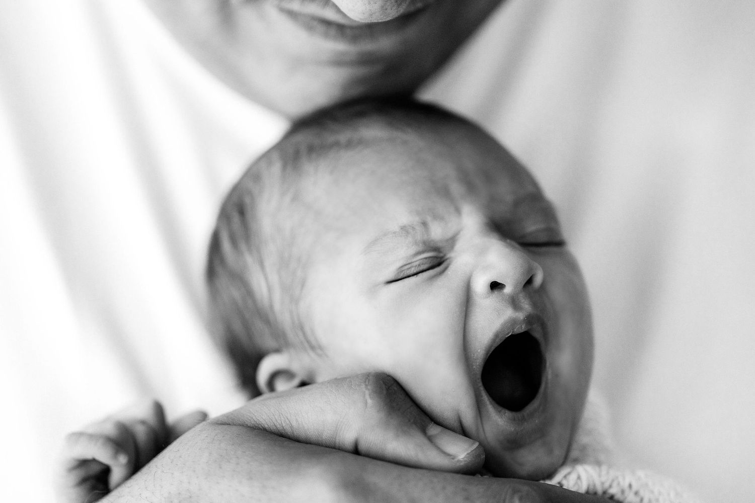Black and white close-up of a yawning newborn baby cradled in a parent's hands.