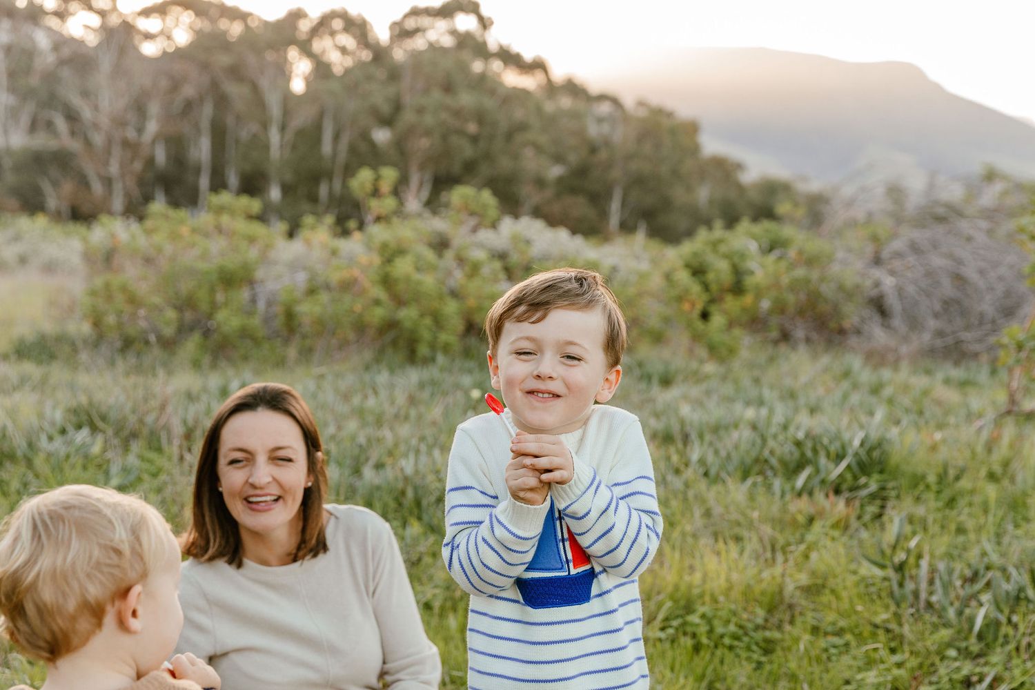 A joyful young boy stands smiling while his mother and baby sibling sit in a scenic outdoor meadow.
