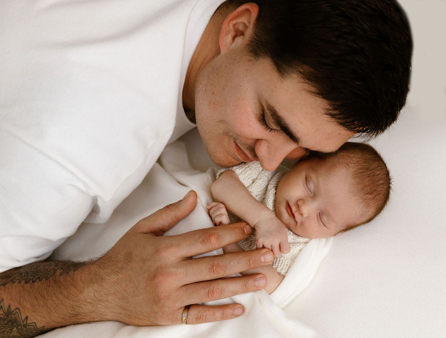Father leans down to kiss sleeping newborn baby lying on white blanket.