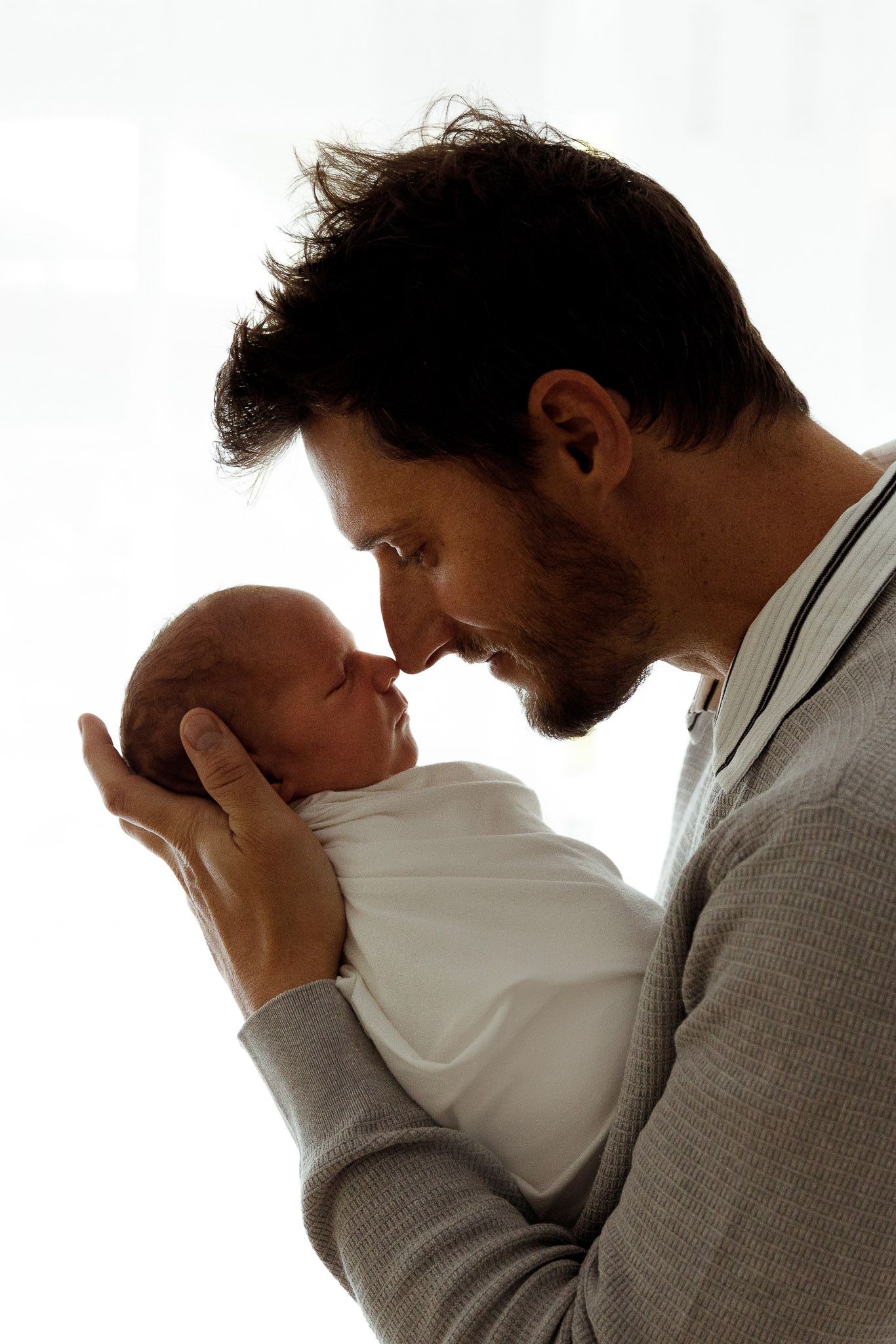 A father tenderly holds his newborn baby wrapped in white, touching foreheads in a loving moment.
