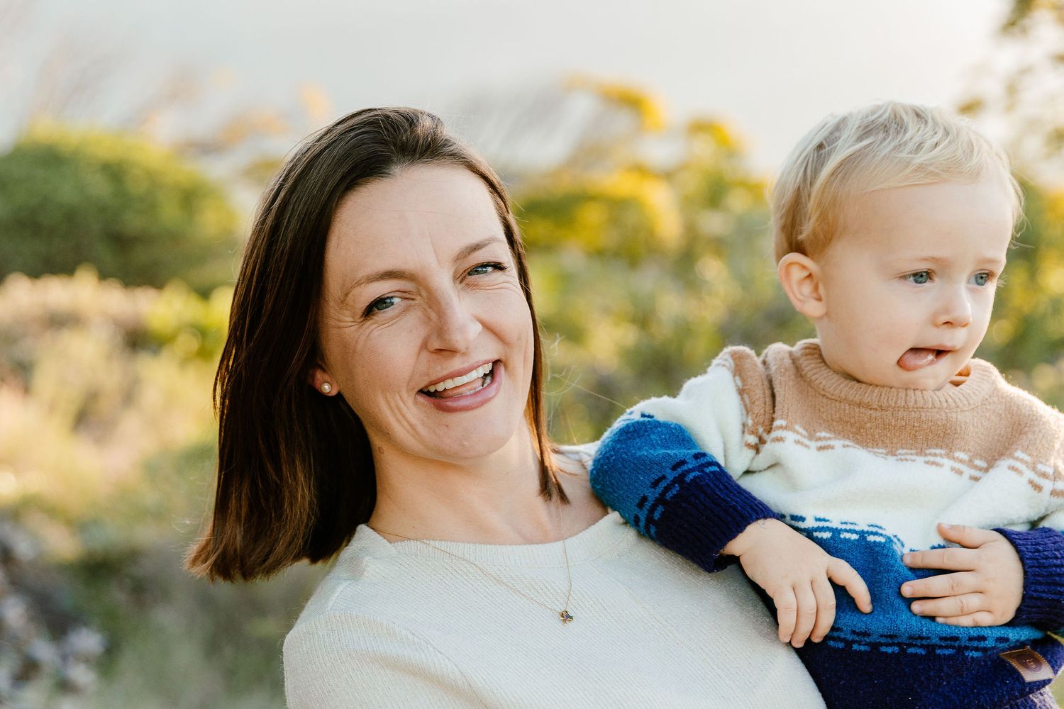 Smiling brunette woman holding a blonde toddler in a blue sweater outdoors in golden light.